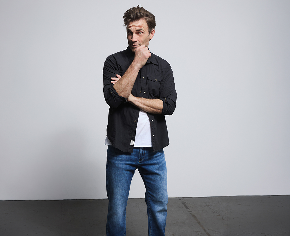 Ein Mann in einem schwarzen Hemd, einem weißen T-Shirt und blauen Jeans steht drinnen auf einem Betonboden, einen Arm verschränkt und die andere Hand am Kinn, und blickt vor einem schlichten, hellen Hintergrund nachdenklich in die Kamera.