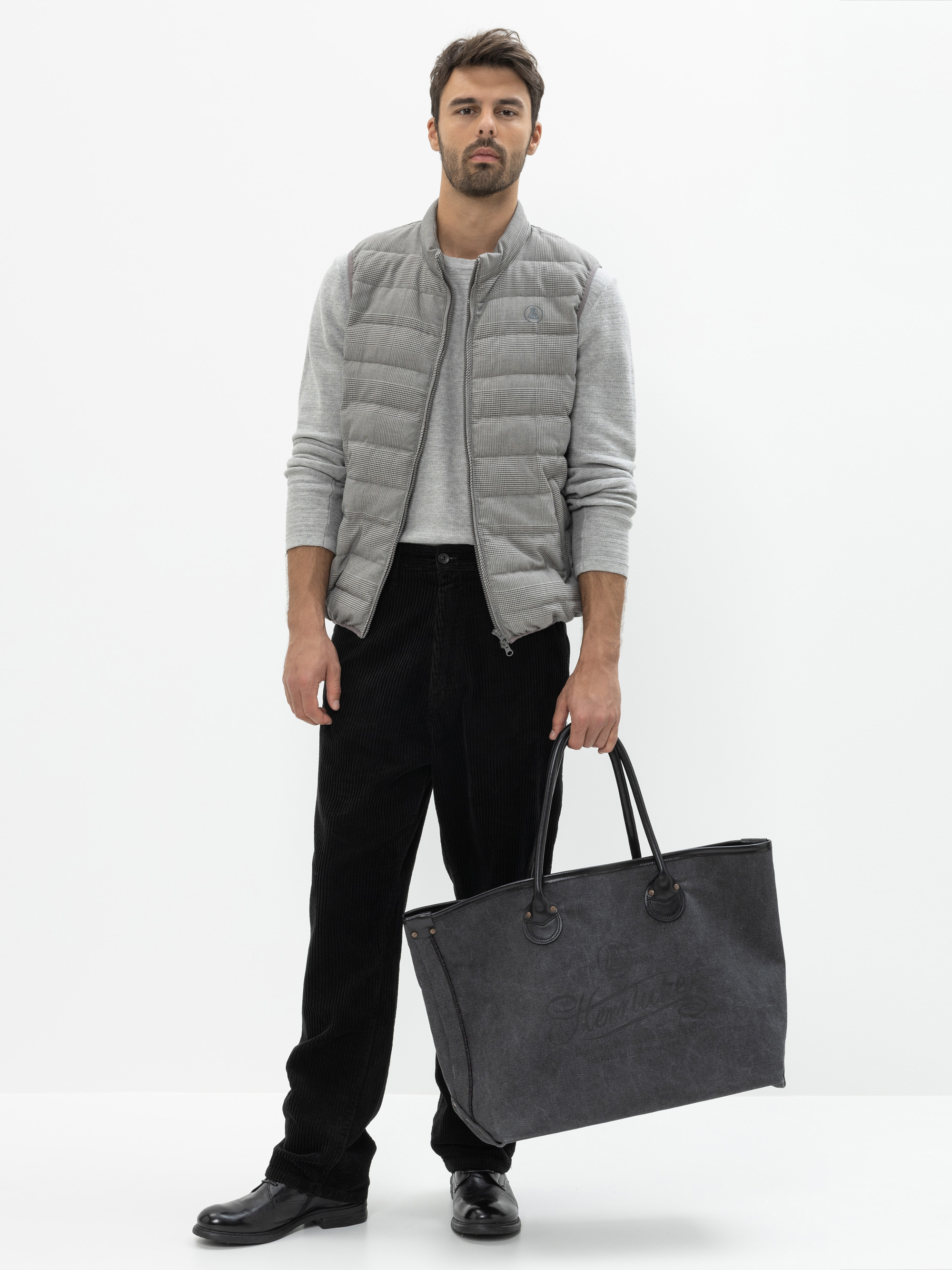 Ein Mann, der eine hellgraue Steppweste über einem langärmeligen Hemd, eine schwarze Hose und schwarze Schuhe trägt, steht vor einem weißen Hintergrund und hält eine große dunkelgraue Tragetasche in seiner linken Hand.