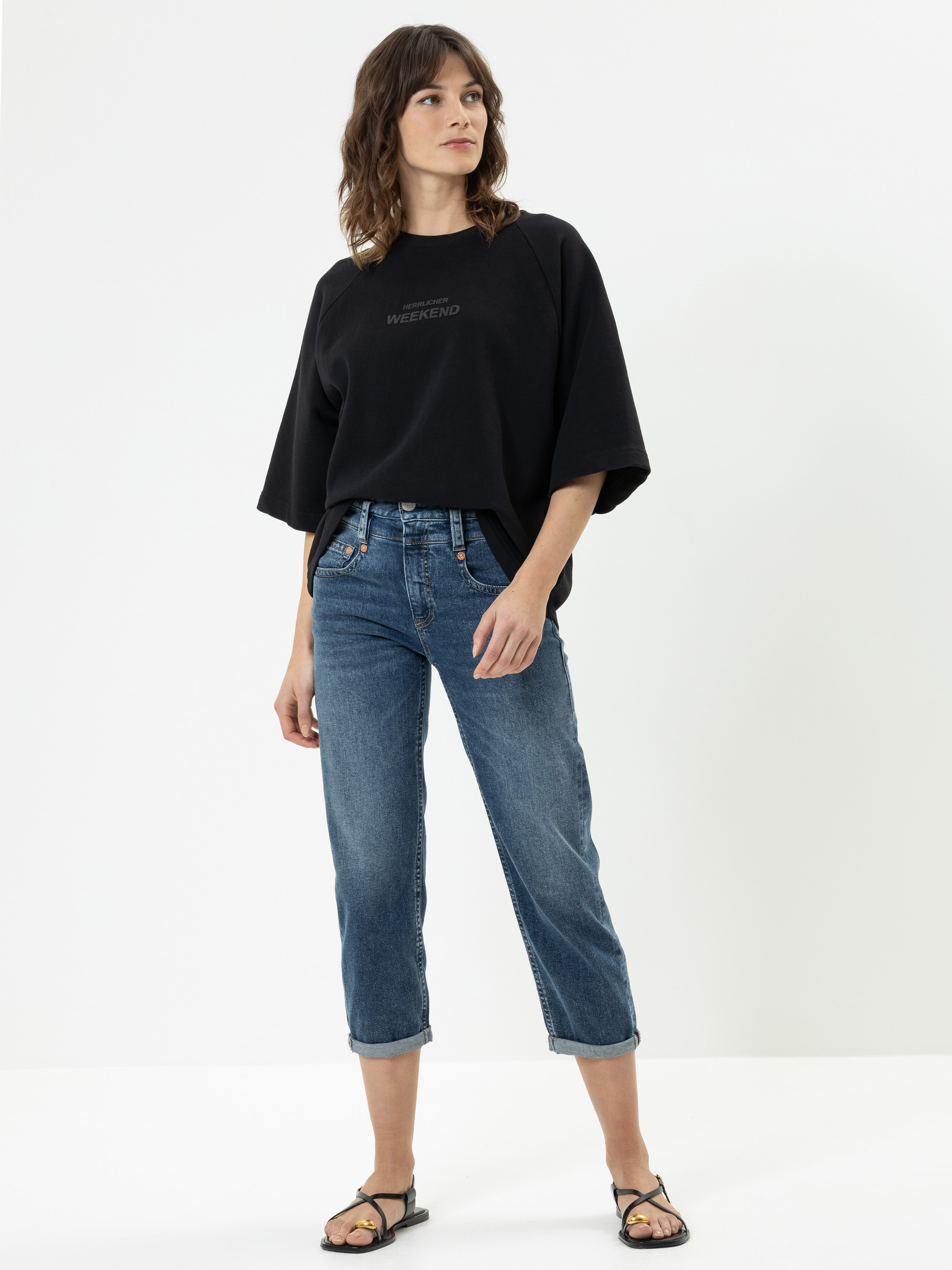 Eine Frau steht vor einem weißen Hintergrund und trägt ein lockeres schwarzes T-Shirt mit dem Aufdruck WEEKEND, eine blaue, kurze Jeans und schwarze Sandalen. Sie hat gewelltes, schulterlanges Haar und ihre Hände stecken in den Taschen.