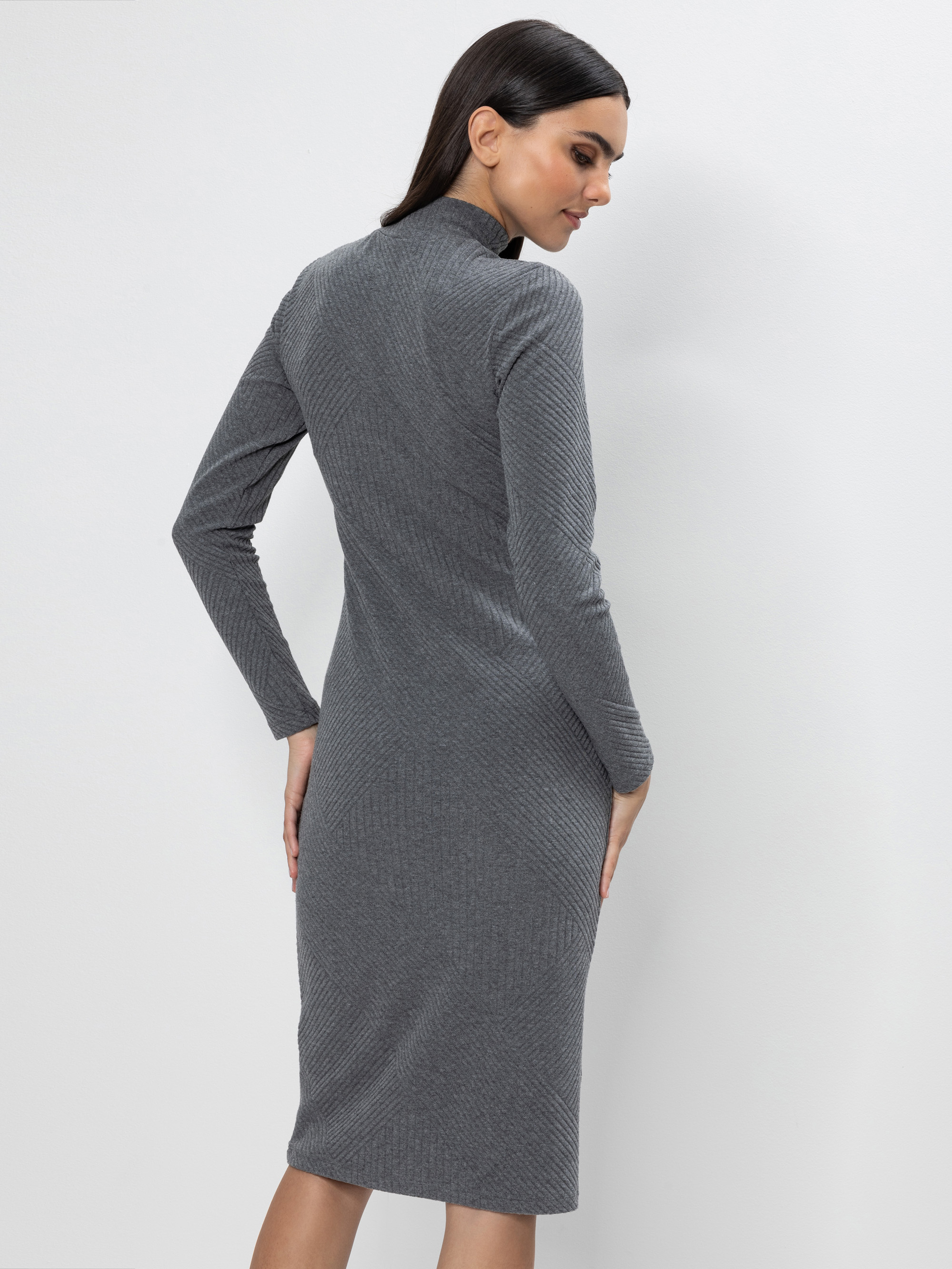 Eine Frau in einem langärmeligen, knielangen, grauen Rippenkleid steht mit dem Gesicht zur Kamera, die rechte Hand auf die Hüfte gestützt, vor einem schlichten, hellen Hintergrund.