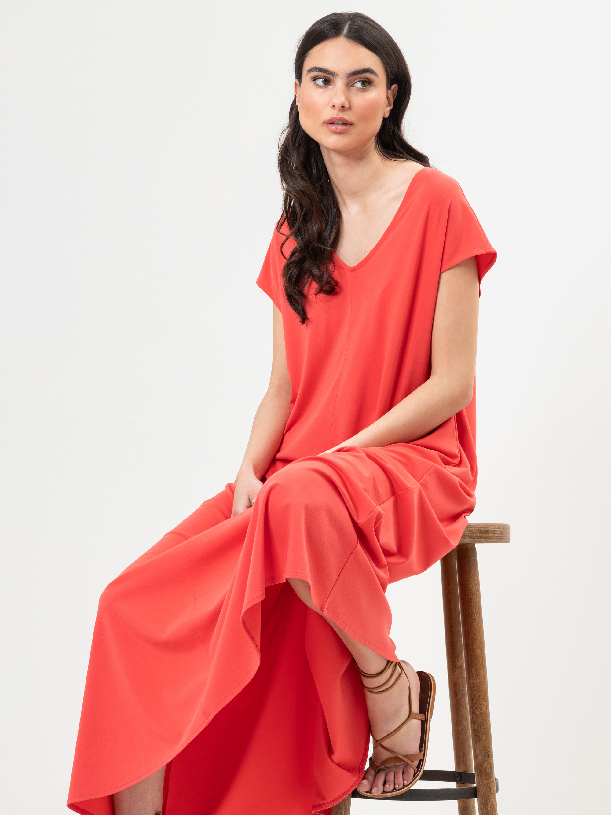 Eine Frau mit langem, dunklem Haar sitzt auf einem Holzschemel, trägt ein locker sitzendes, leuchtend korallenrotes Maxikleid und braune Riemchensandalen vor einem schlichten weißen Hintergrund.