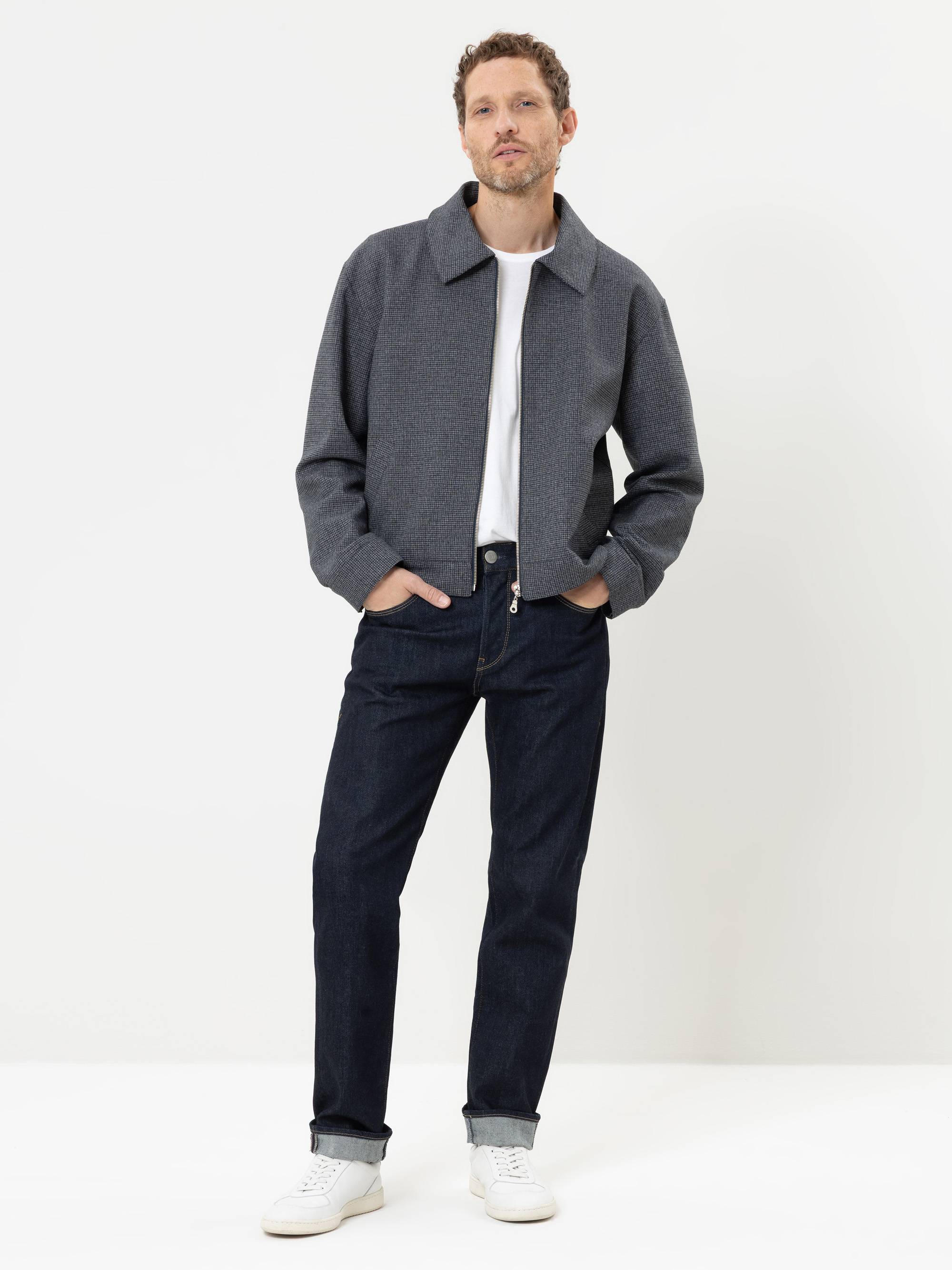 Vor einem weißen Hintergrund steht ein Mann, der eine graue Jacke, ein weißes T-Shirt, dunkelblaue Jeans und weiße Turnschuhe trägt; er hat die Hände in den Taschen und lächelt leicht.
