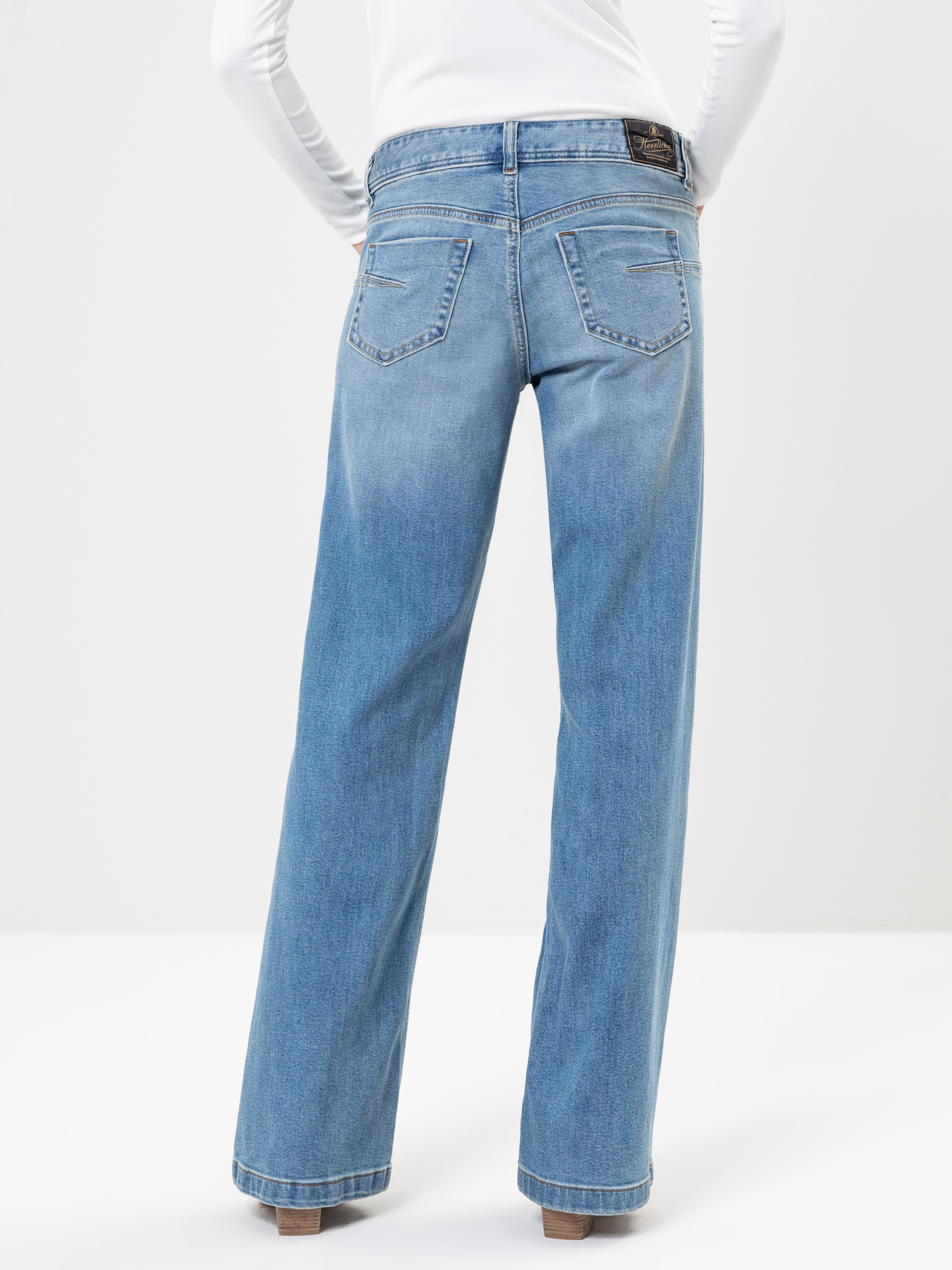 Eine Person, die hellblaue, hochtaillierte Jeans mit weitem Bein und ein weißes langärmeliges Oberteil trägt, von hinten vor einem einfarbig weißen Hintergrund dargestellt.