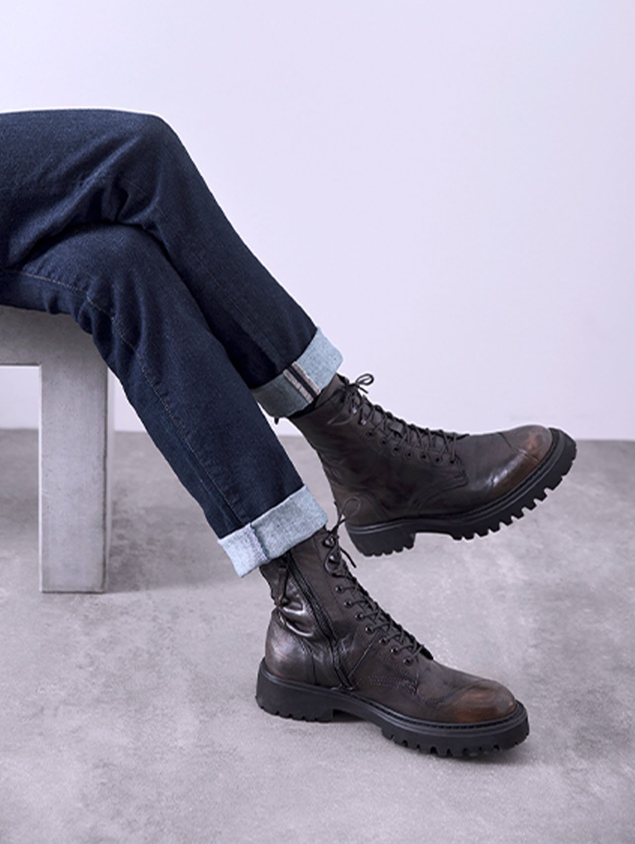 Eine Person sitzt auf einer Betonbank und trägt dunkelblaue, hochgekrempelte Jeans und dunkelbraune, geschnürte Lederstiefel mit rauen Sohlen, die vor einem hellgrauen Hintergrund gezeigt werden.