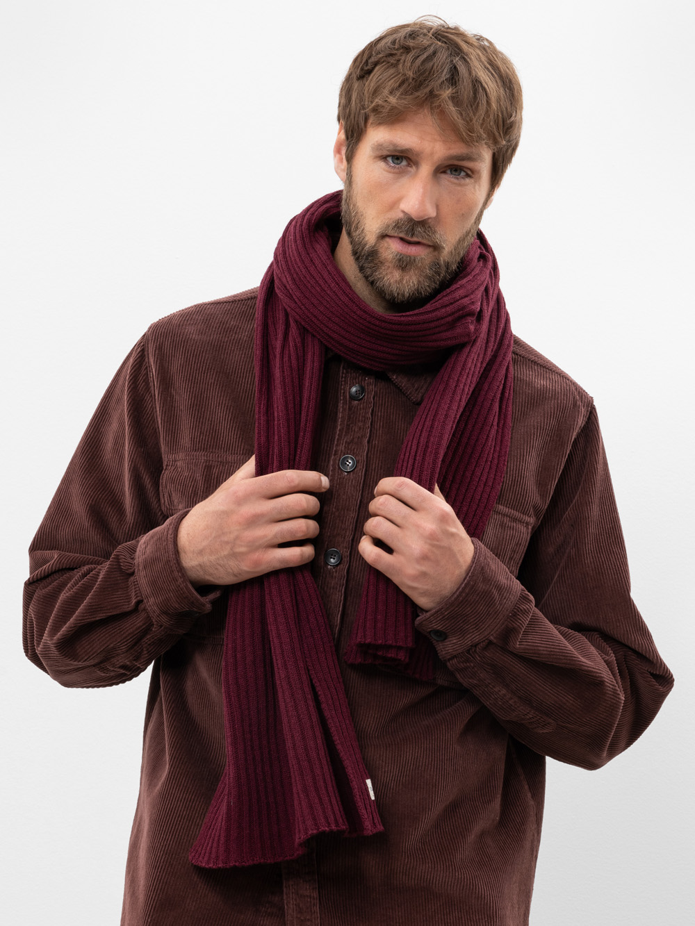 Ein Mann mit hellbraunem Haar und Bart trägt einen kastanienbraunen Schal und ein braunes Cordhemd zum Aufknöpfen und steht vor einem schlichten weißen Hintergrund. Er schaut in die Kamera, während er seinen Schal zurechtrückt.