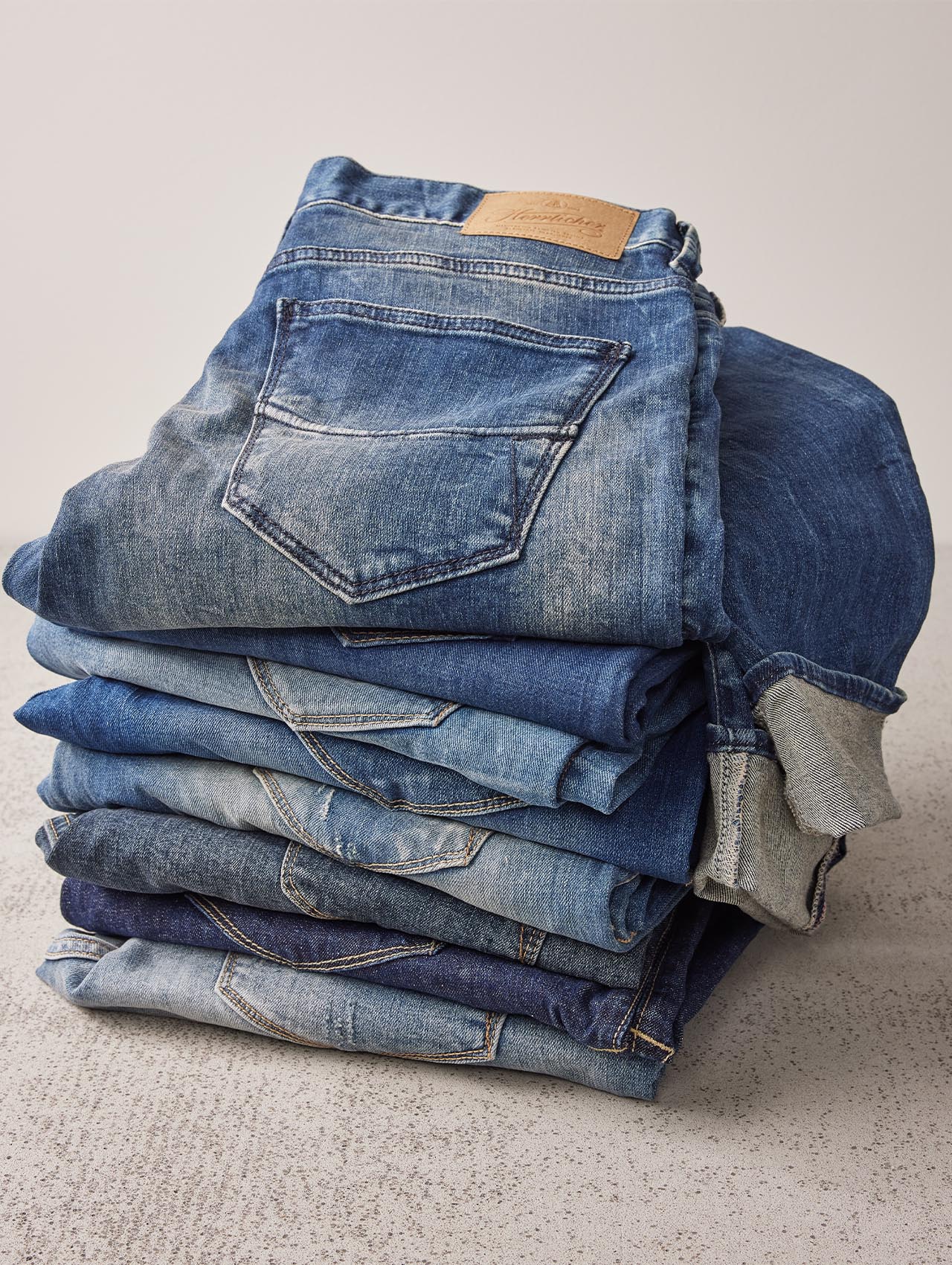 Ein ordentlich gefalteter Stapel blauer Jeans in verschiedenen Farbtönen liegt auf einer hellen, strukturierten Oberfläche, wobei das oberste Paar eine sichtbare Gesäßtasche und einen hellbraunen Lederaufnäher aufweist.