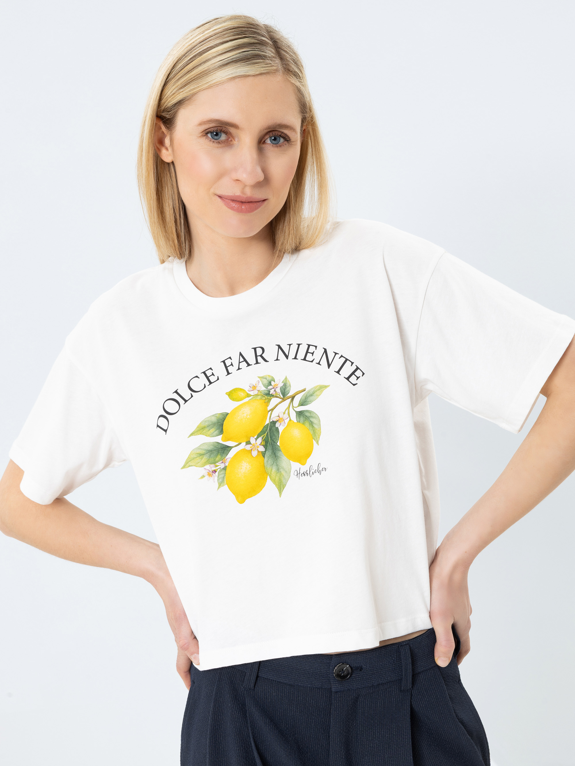 Eine Frau mit blonden Haaren, die ein weißes T-Shirt mit der Aufschrift DOLCE FAR NIENTE und einer Illustration von Zitronen und grünen Blättern trägt, steht mit den Händen in den Hüften und lächelt sanft.
