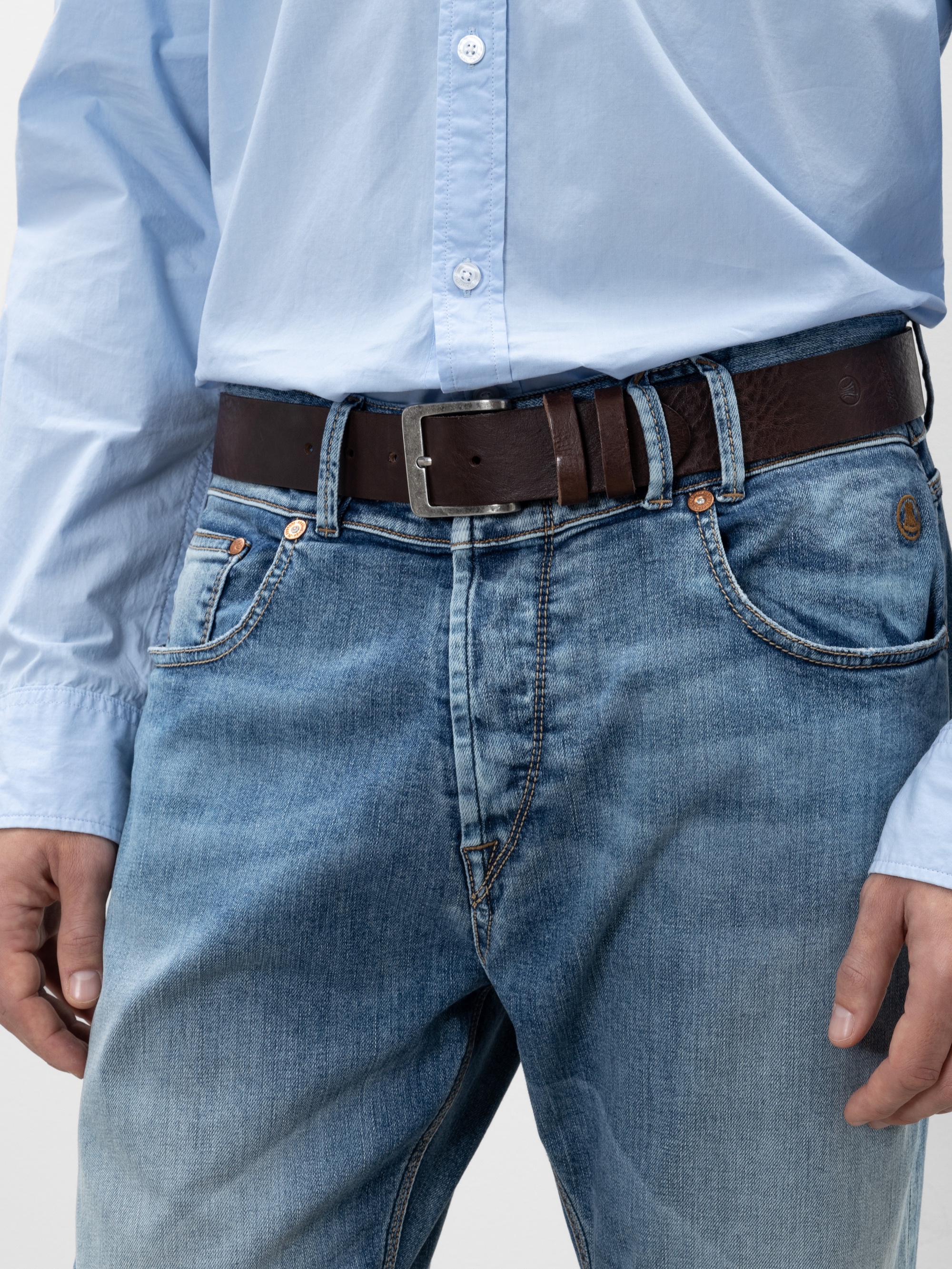Eine Person, die ein hellblaues Button-up-Hemd trägt, das in eine blaue Jeans mit einem dunkelbraunen Ledergürtel gesteckt ist; nur der Oberkörper und die Oberschenkel sind sichtbar.