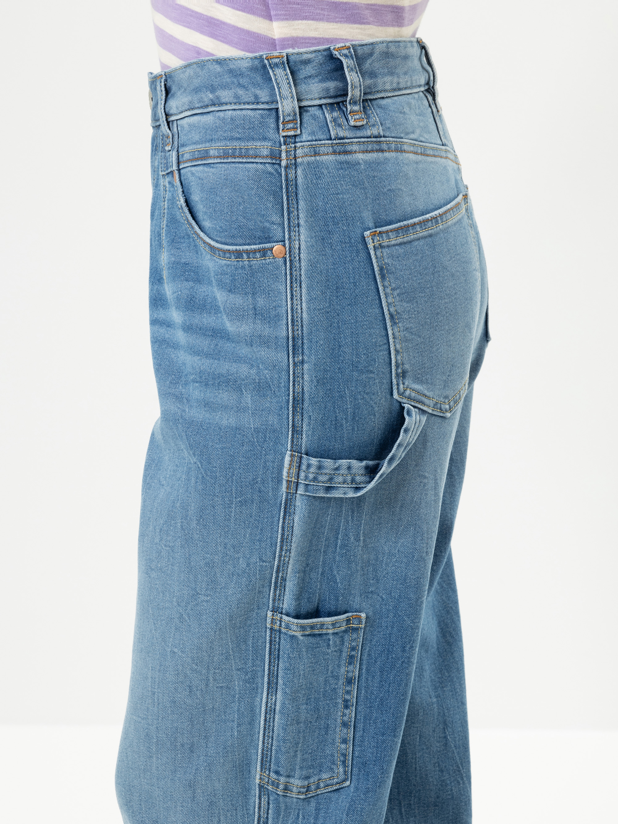 Eine Person trägt eine hellblaue Jeans mit hoher Taille, mehreren Taschen und einer Hammerschlaufe, von der Seite gesehen. Die Person trägt außerdem ein hellviolett und weiß gestreiftes Hemd, das in die Hose gesteckt ist.