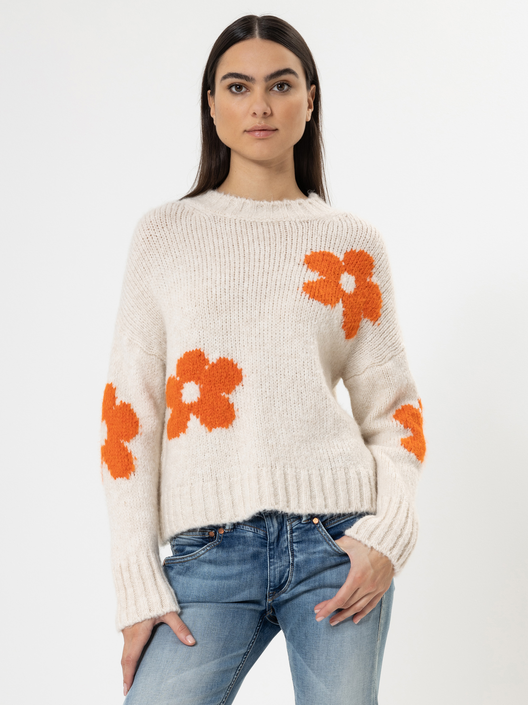 Eine Frau, die einen cremefarbenen Pullover mit orangefarbenen Blumenmustern trägt, steht vor einem weißen Hintergrund. Sie hat langes dunkles Haar, trägt eine blaue Jeans und hat eine Hand in der Tasche.
