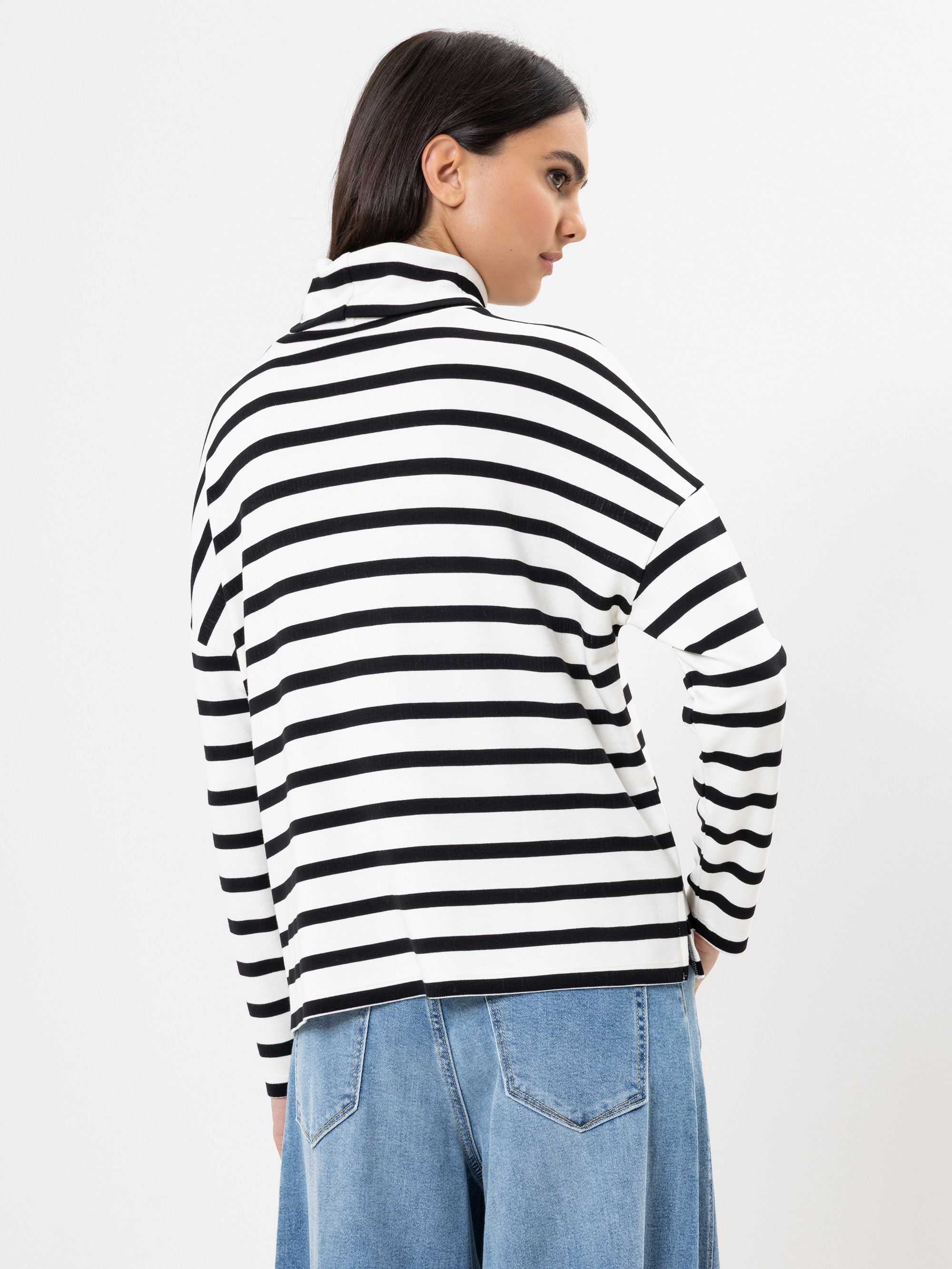 Eine Frau mit glattem dunklem Haar trägt ein schwarz-weiß gestreiftes langärmeliges Oberteil und eine blaue Jeans. Sie blickt von der Kamera weg auf einen schlichten weißen Hintergrund.