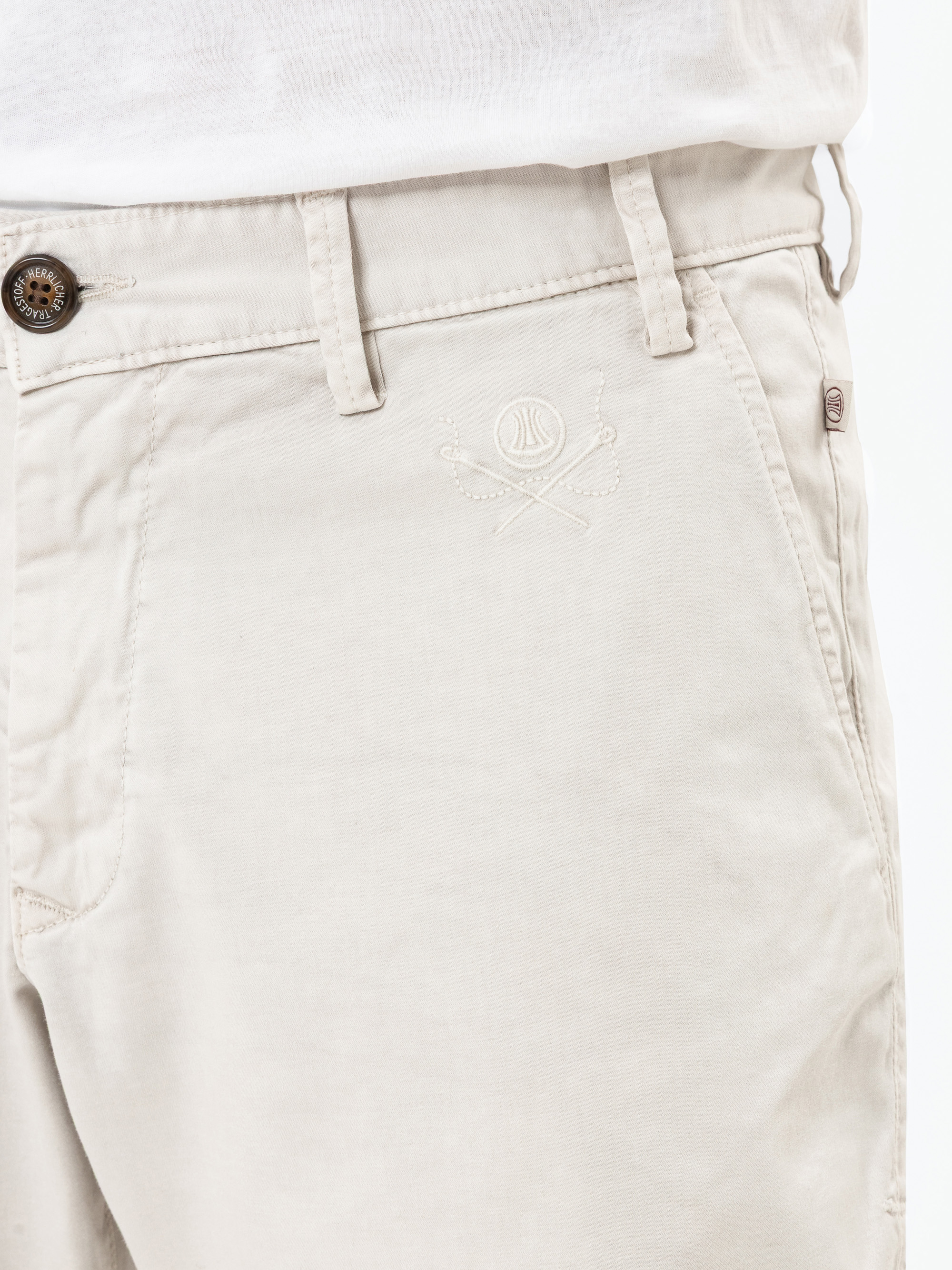 Nahaufnahme einer Person, die eine hellbeige Hose mit Knopfverschluss, Gürtelschlaufen und einem gestickten Logo in der Nähe der Tasche trägt, kombiniert mit einem weißen Hemd.