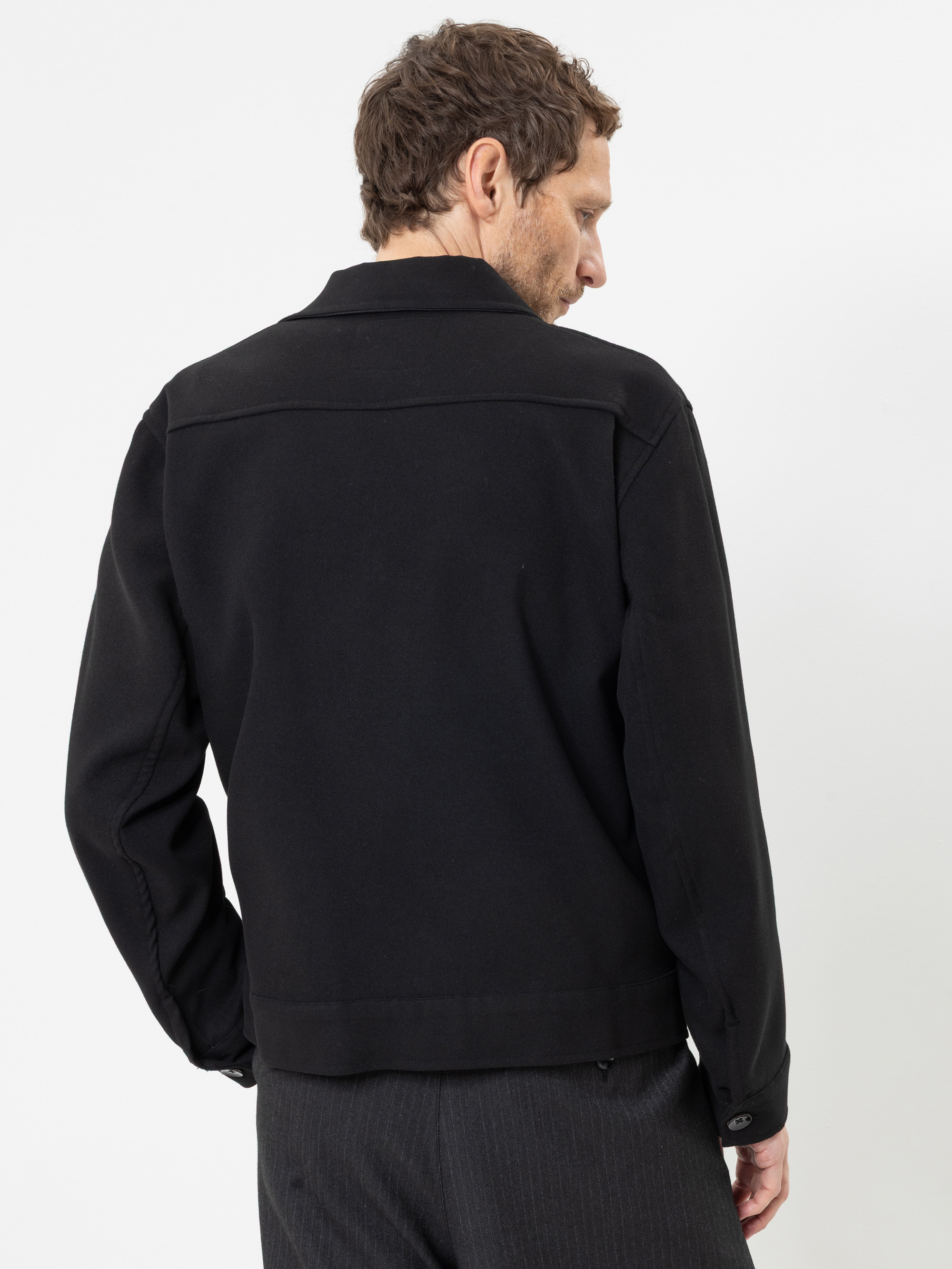 Ein Mann mit kurzen braunen Haaren, der eine schwarze Jacke mit Kragen und eine dunkle Hose trägt, steht mit dem Rücken zur Kamera, den Kopf leicht zur Seite gedreht, vor einem schlichten weißen Hintergrund.