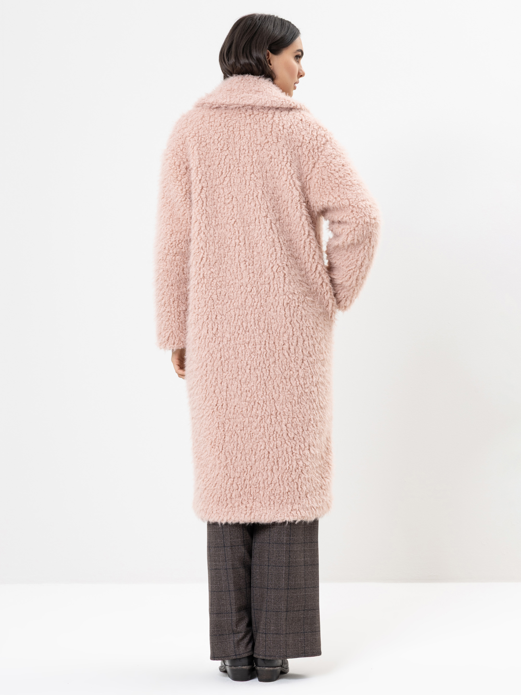 Eine Frau steht mit dem Gesicht zur Wand, sie trägt einen langen, blassrosa, wuscheligen Mantel über einer grau-karierten Hose und schwarzen Schuhen, vor einem schlichten weißen Hintergrund.