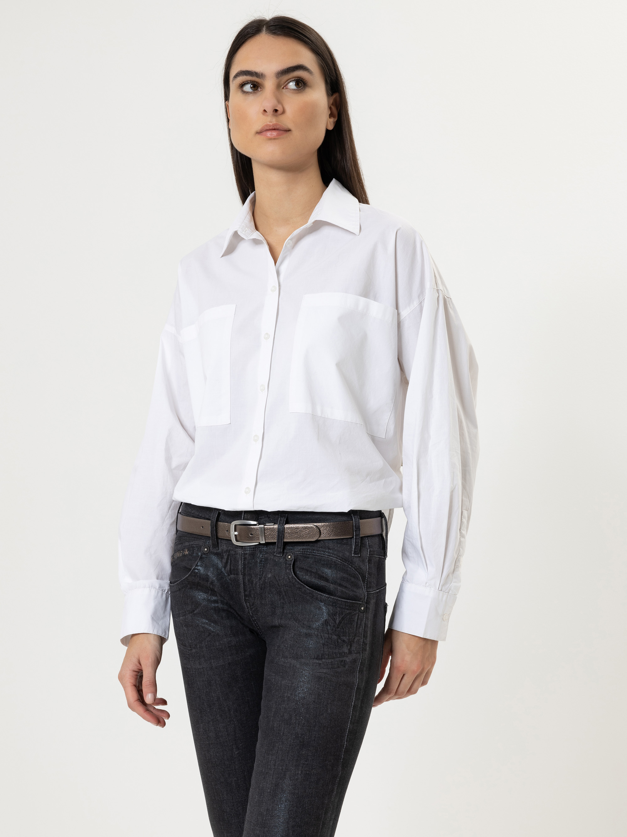 Eine Frau mit langen dunklen Haaren trägt ein weißes Button-up-Hemd mit Vordertaschen, schwarze Jeans und einen schwarzen Gürtel. Sie steht vor einem schlichten hellen Hintergrund und blickt leicht zur Seite.