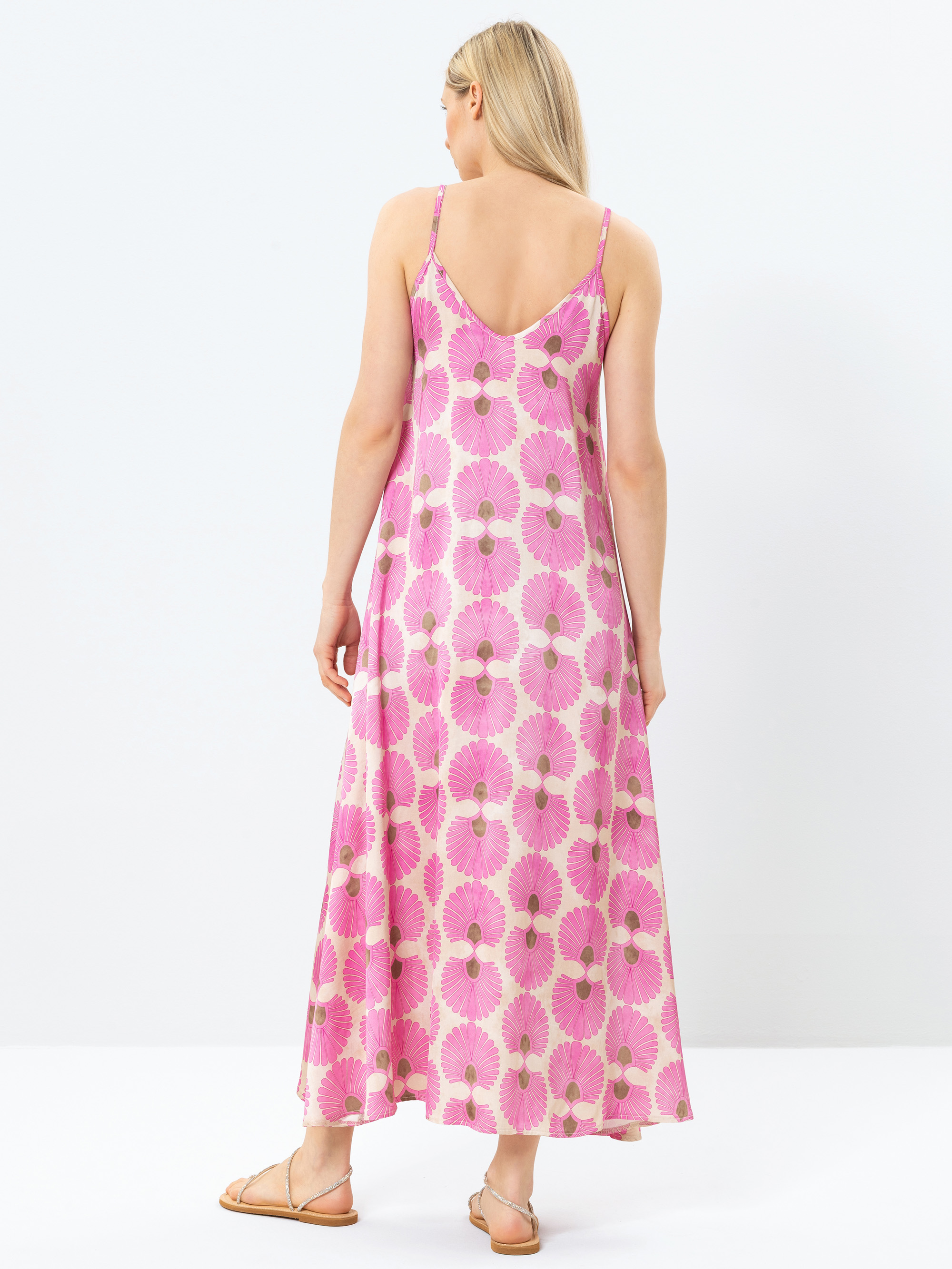 Eine Frau mit langen blonden Haaren steht mit dem Gesicht zur Wand und trägt ein ärmelloses Maxikleid mit dünnen Trägern und flachen Sandalen mit rosa und weißem Blumenmuster vor einem schlichten weißen Hintergrund.