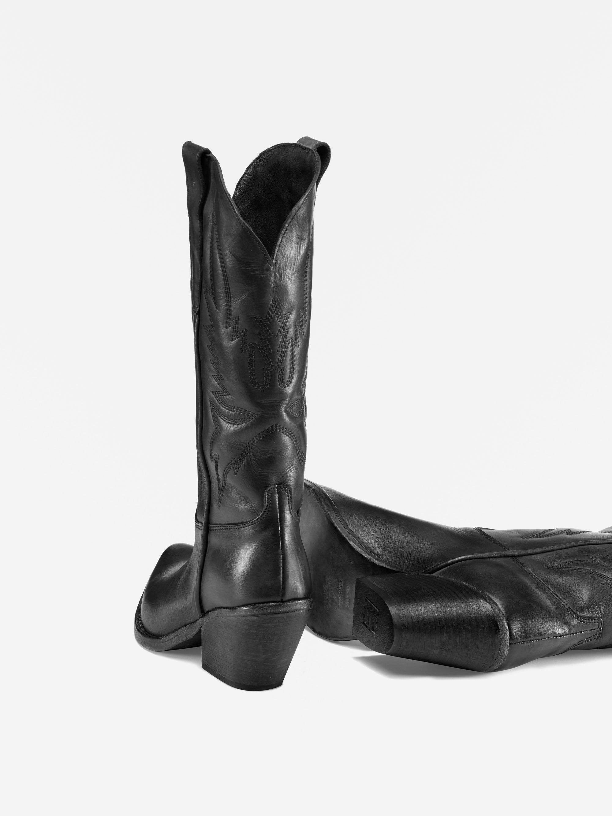 Ein Paar hohe Cowboystiefel aus schwarzem Leder mit Ziernähten, wobei ein Stiefel aufrecht steht und der andere auf der Seite liegt, dargestellt vor einem weißen Hintergrund.