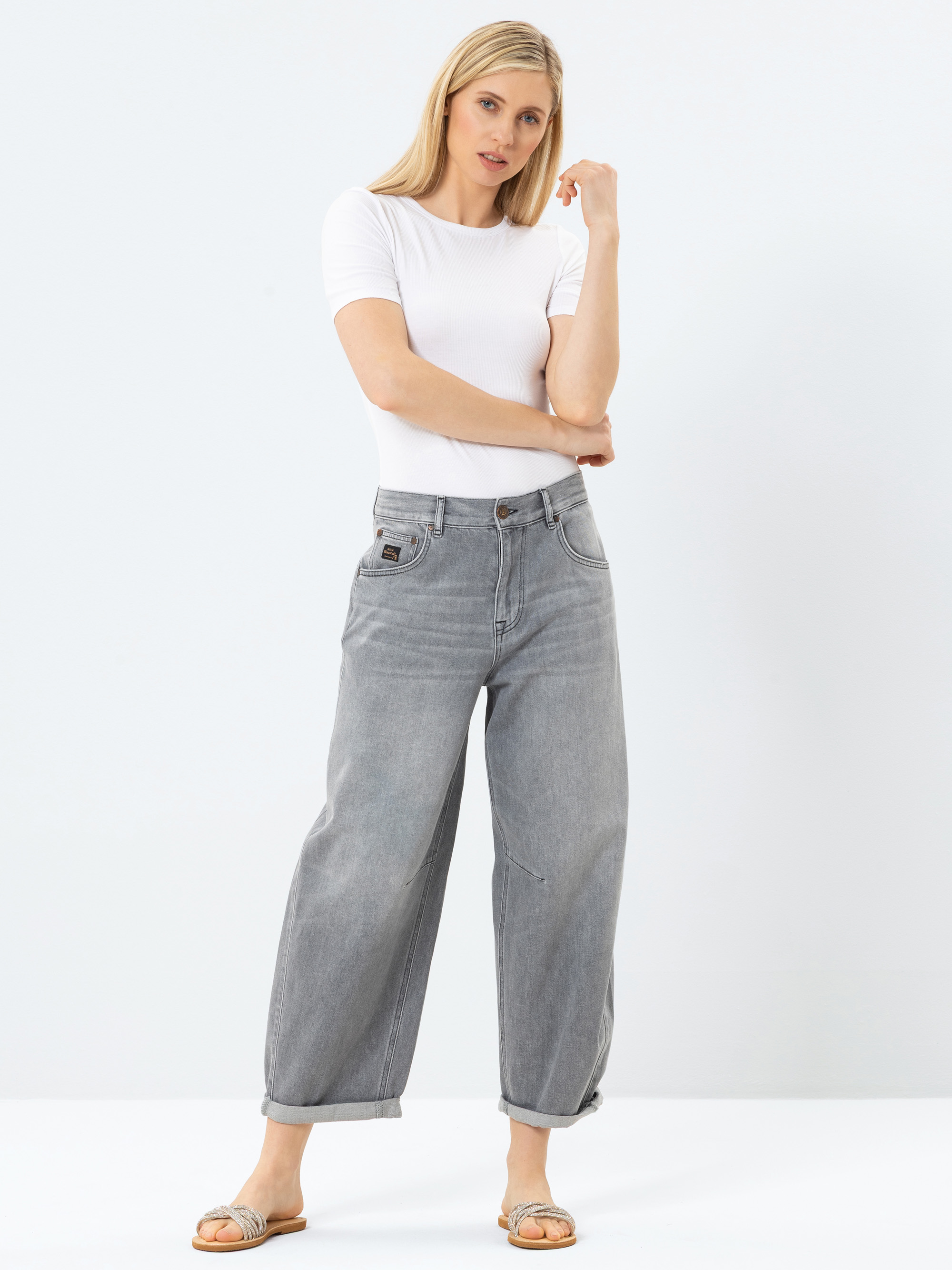 Eine Frau mit langen blonden Haaren trägt ein weißes T-Shirt, eine weite graue Jeans mit aufgerollten Manschetten und beigefarbene Slide-Sandalen, die vor einem schlichten weißen Hintergrund stehen.