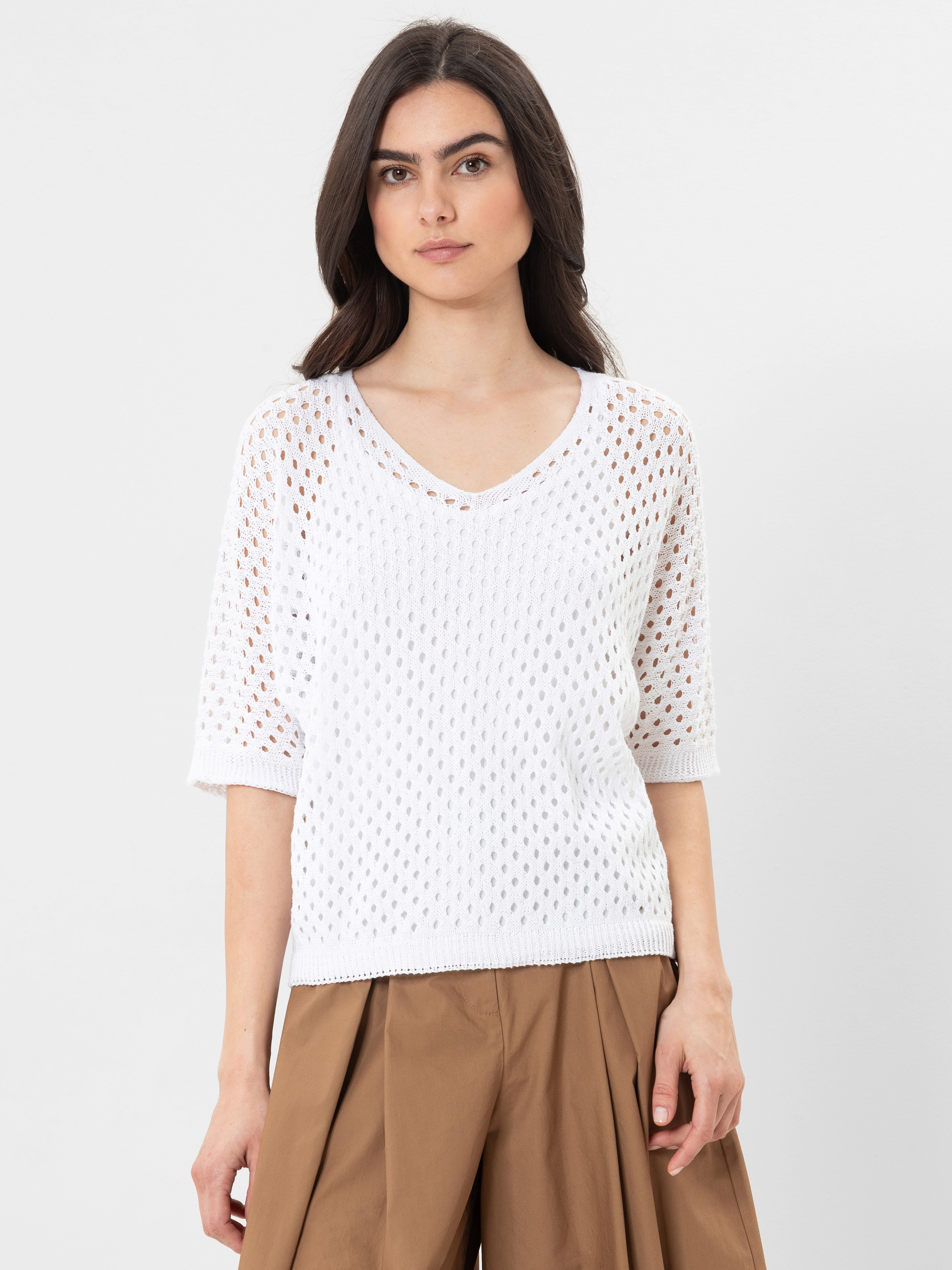Eine Frau mit langen dunklen Haaren, die einen weißen, kurzärmeligen Strickpullover mit V-Ausschnitt und eine braune Bundfaltenhose trägt, steht vor einem schlichten weißen Hintergrund.