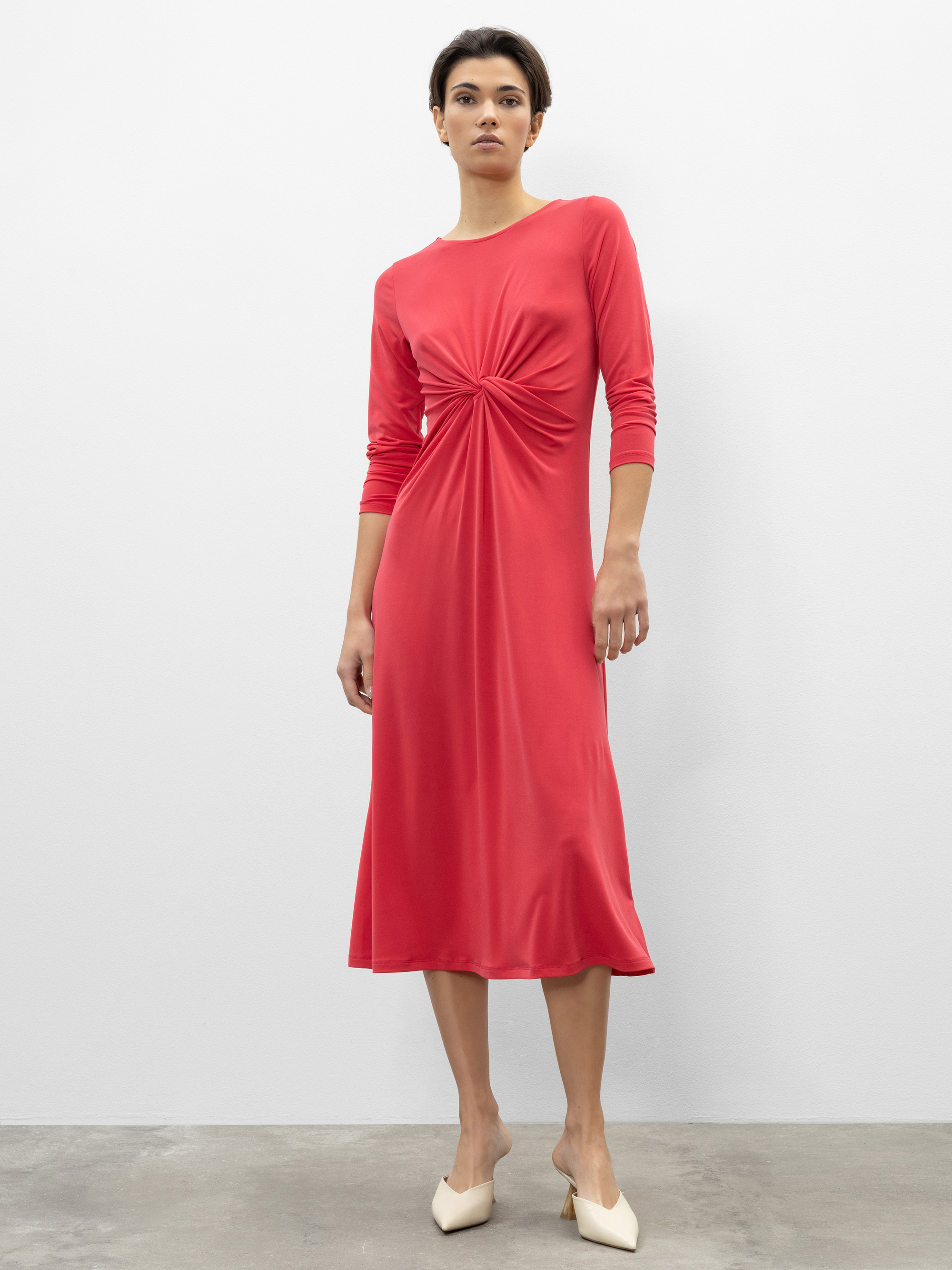 Eine Person steht vor einer schlichten weißen Wand und trägt ein langärmeliges, halblanges rotes Kleid mit einem gerafften Knoten in der Taille und weißen spitzen Schuhen.