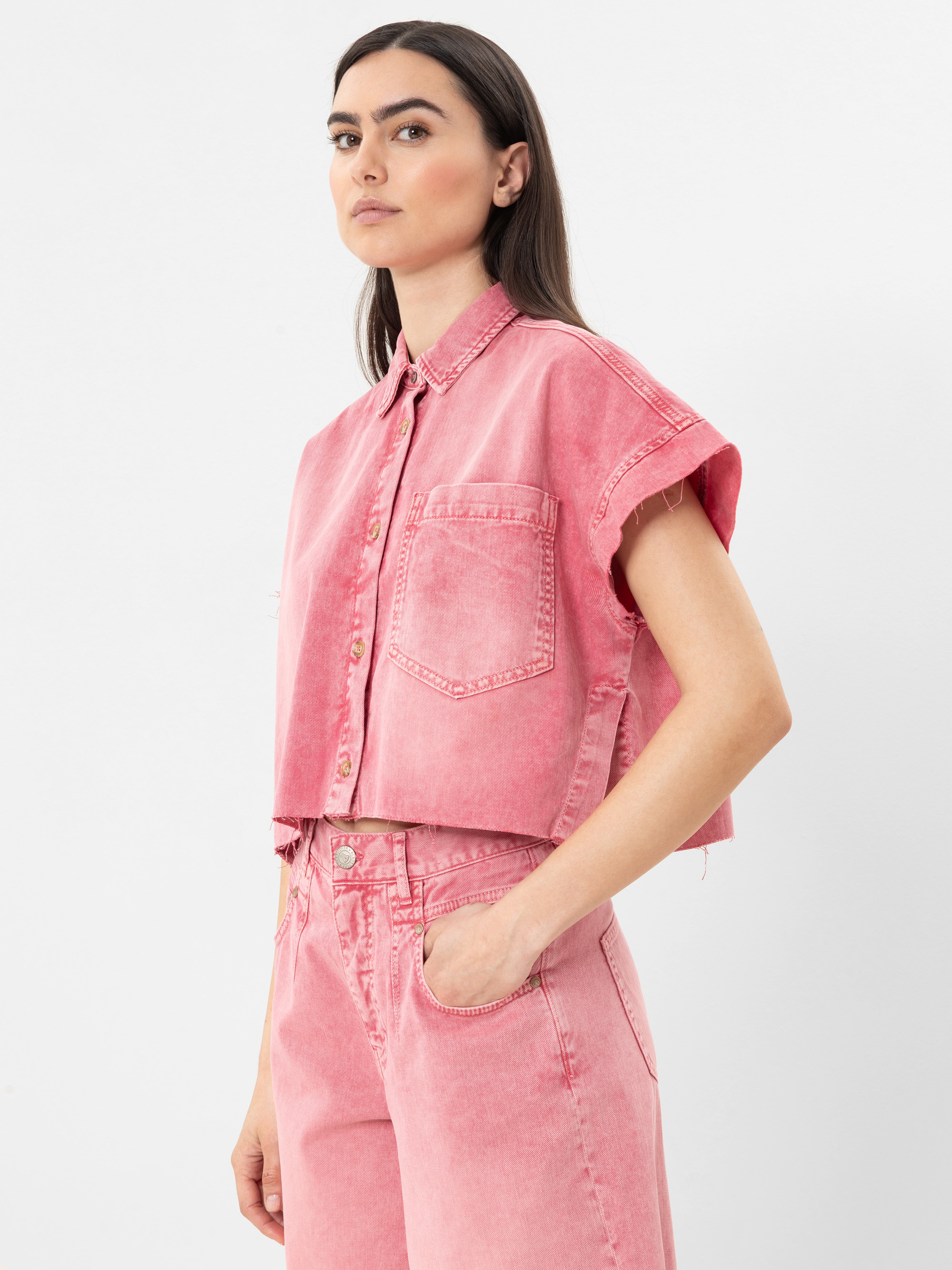 Eine Frau steht vor einem weißen Hintergrund und trägt ein kurzärmeliges, pinkfarbenes Jeanshemd zum Aufknöpfen und dazu passende Jeans mit hoher Taille. Sie hat langes braunes Haar, eine Hand in der Tasche und blickt selbstbewusst zur Seite.