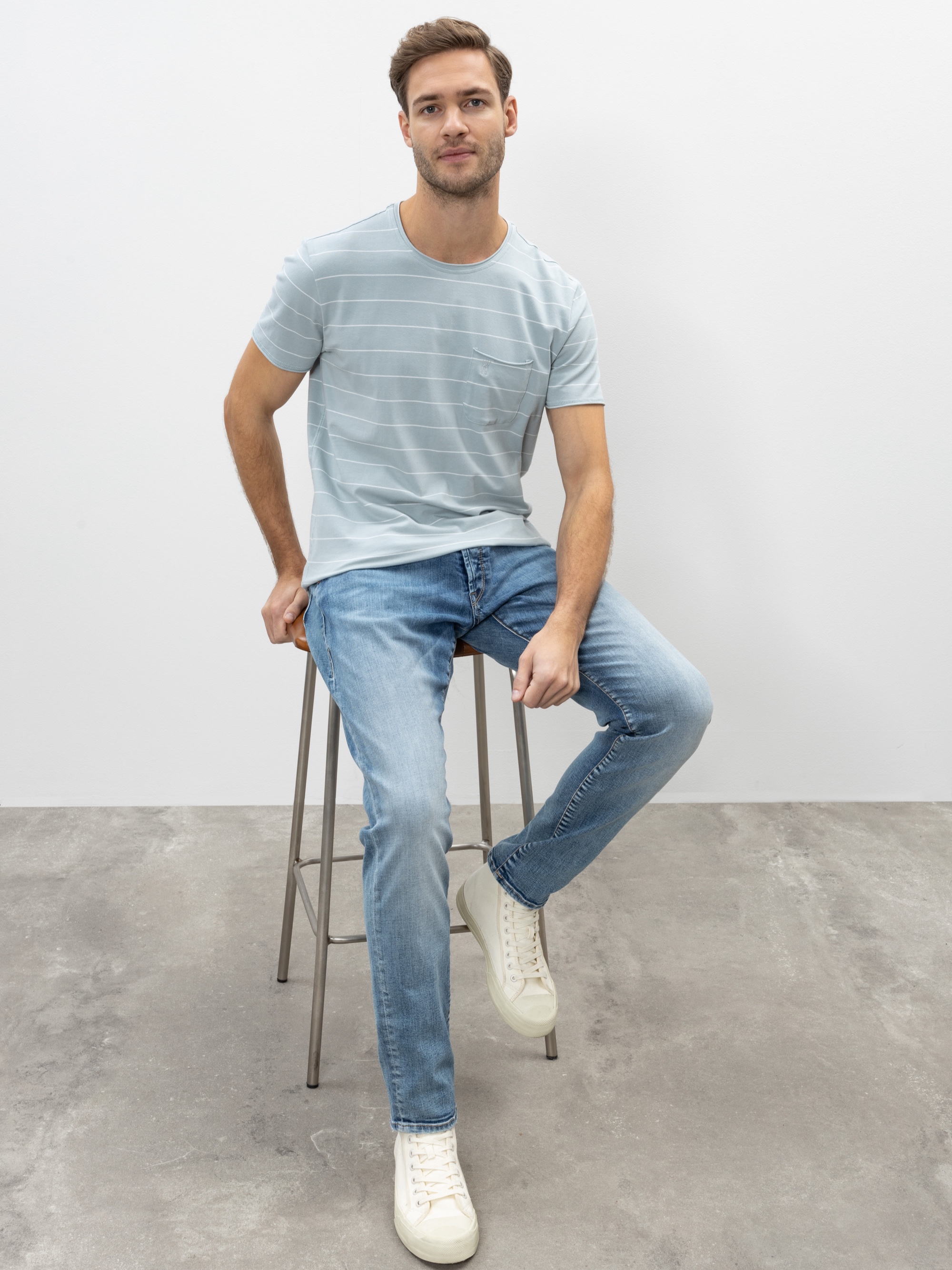 Ein Mann mit kurzen braunen Haaren in einem hellblau gestreiften T-Shirt, blauen Jeans und weißen Turnschuhen sitzt auf einem Metallhocker vor einem schlichten, hellen Hintergrund.