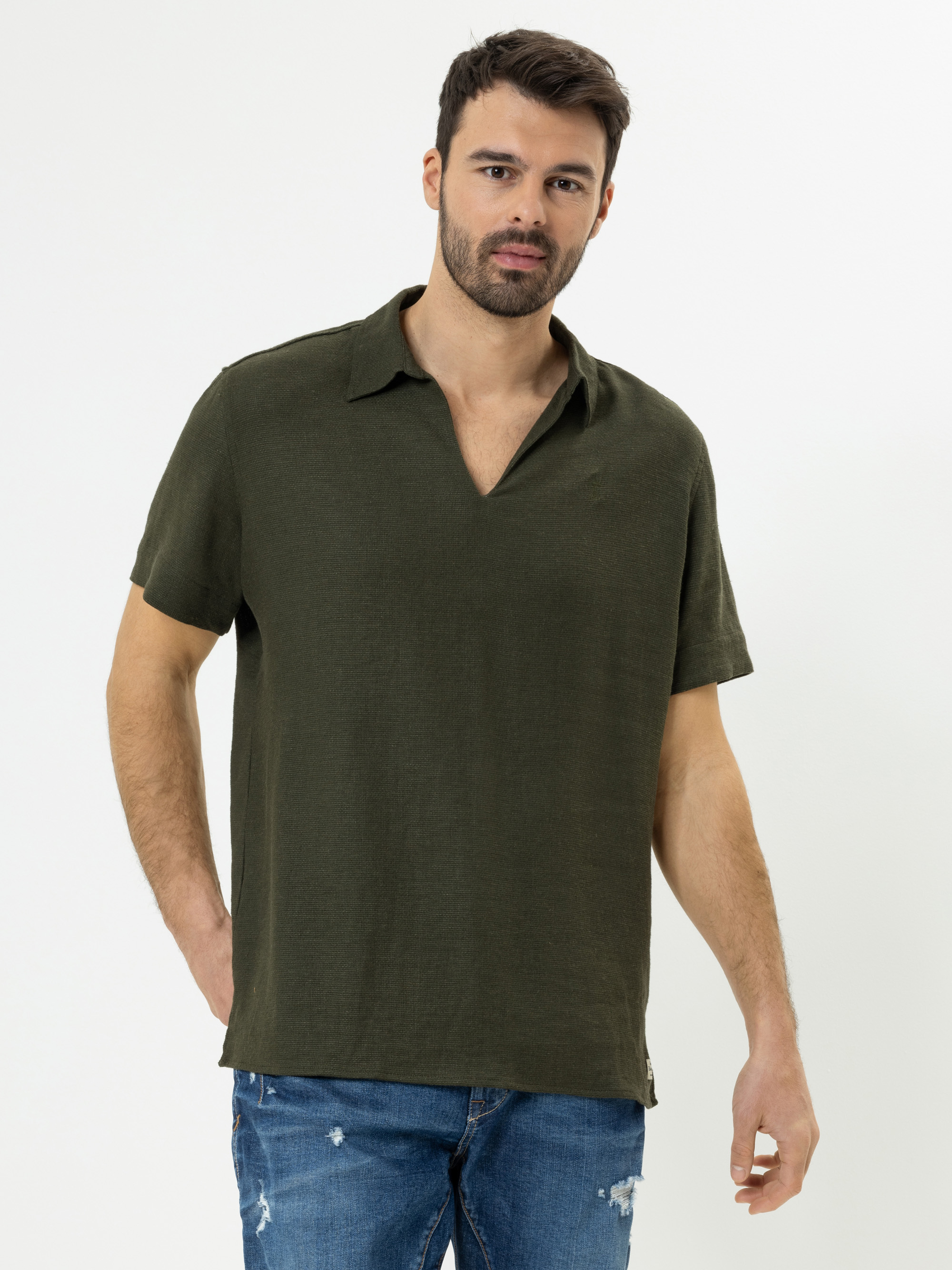 Ein Mann mit kurzen dunklen Haaren und einem Bart trägt ein olivgrünes, kurzärmeliges Hemd mit Kragen und blaue, abgenutzte Jeans und steht vor einem schlichten weißen Hintergrund.