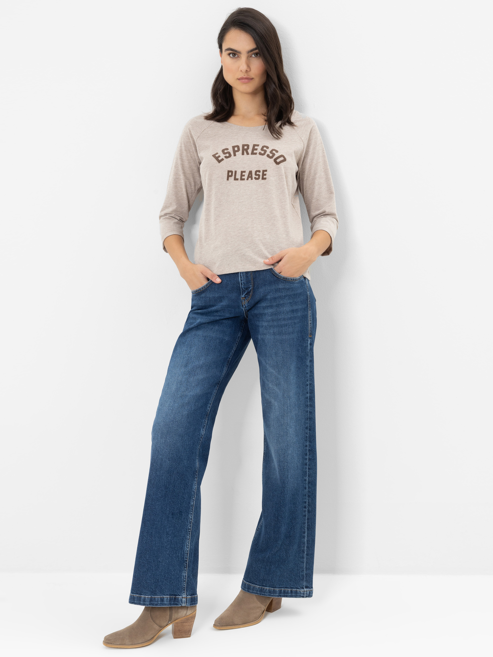 Eine Frau steht vor einer weißen Wand und trägt ein beiges Shirt mit der Aufschrift „ESPRESSO PLEASE“, eine blaue Jeans mit weitem Bein und beige Stiefel mit Absatz. Sie hat eine Hand in der Tasche und sieht entspannt aus.