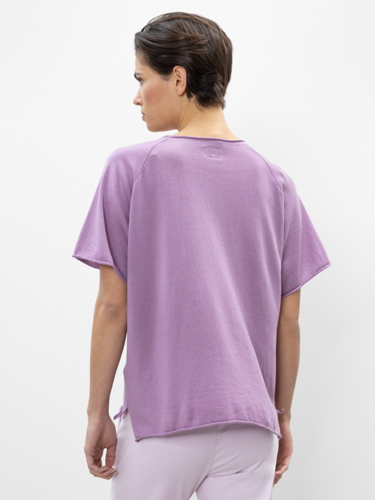 Eine Person mit kurzen braunen Haaren wird von hinten gezeigt, die ein locker sitzendes, kurzärmeliges lavendelfarbenes Oberteil und eine hellviolette Hose trägt und vor einem schlichten weißen Hintergrund steht.