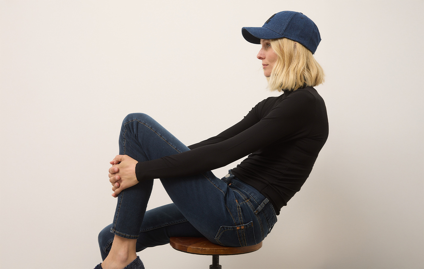 Eine Frau, die ein schwarzes, langärmeliges Oberteil, blaue Jeans und eine blaue Jeansmütze trägt, sitzt seitlich auf einem Hocker vor einem schlichten weißen Hintergrund, die Knie angewinkelt und die Arme auf die Beine gestützt.