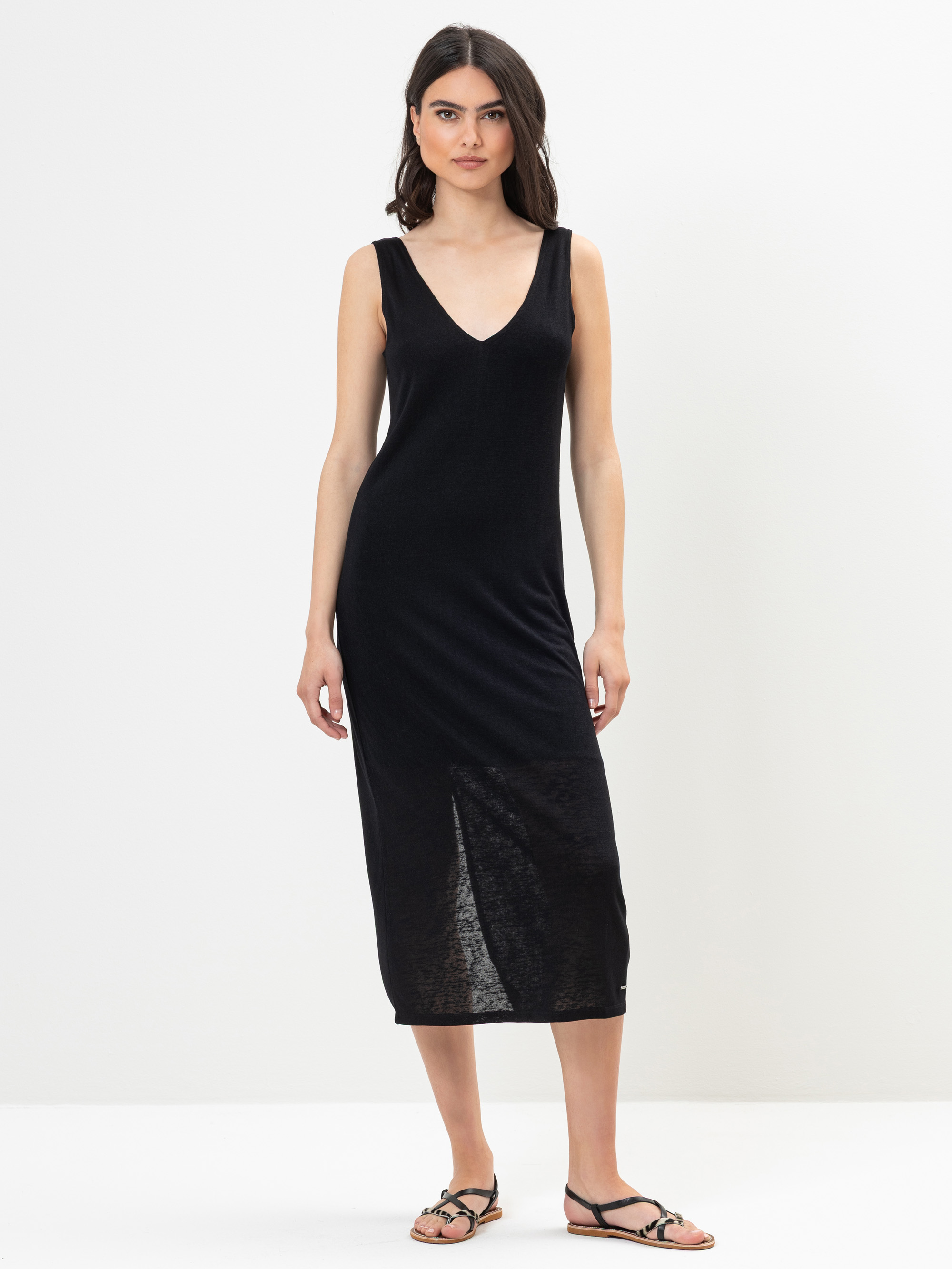 Eine Frau steht vor einem schlichten weißen Hintergrund und trägt ein ärmelloses, schwarzes Midikleid mit V-Ausschnitt und schwarze flache Riemchensandalen. Sie hat langes dunkles Haar und einen neutralen Gesichtsausdruck, wobei sie die Arme entspannt