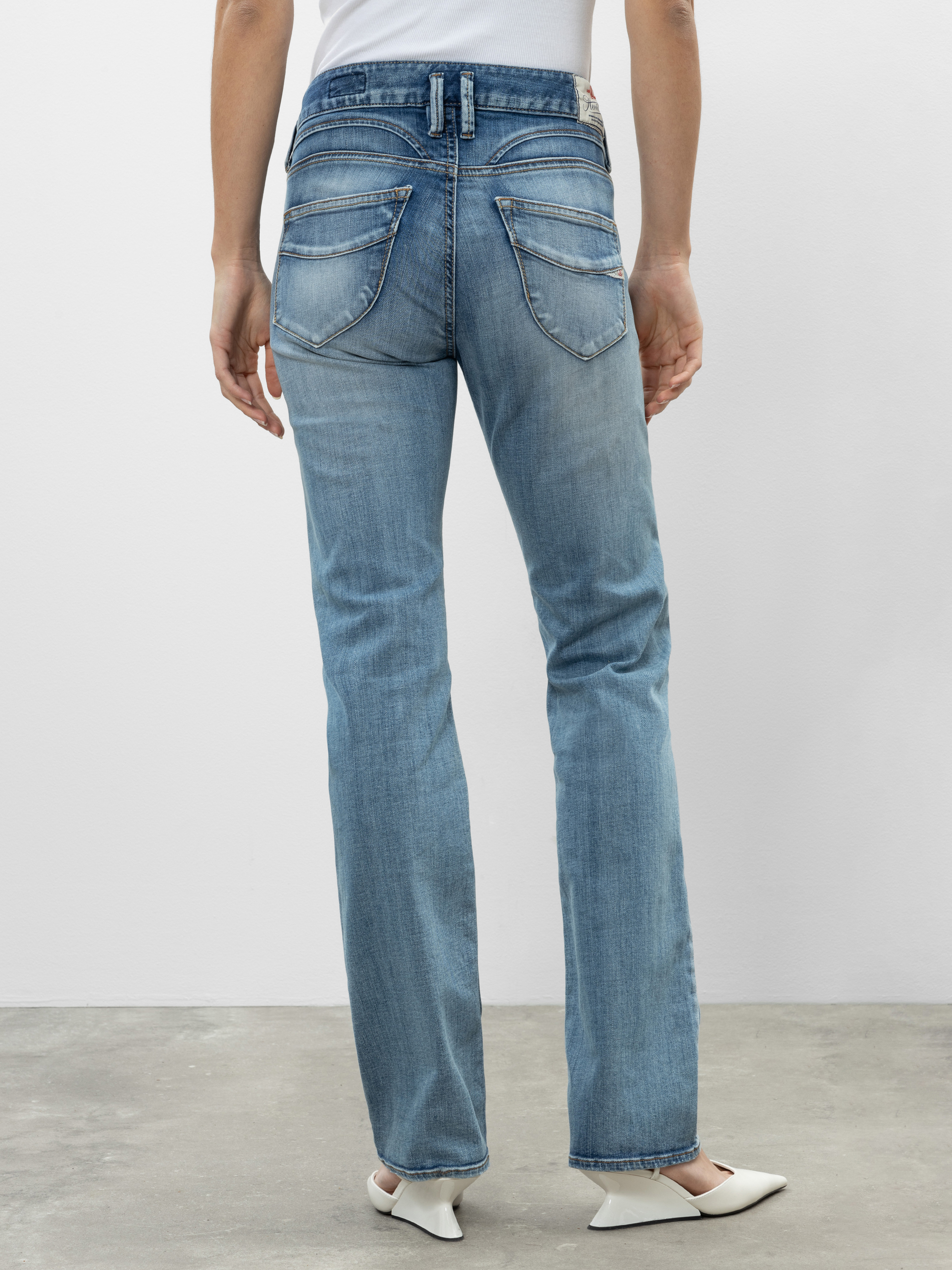 Eine Person steht mit dem Gesicht zur Wand und trägt eine hellblaue Jeans mit geraden Beinen und Gesäßtaschen sowie ein weißes Oberteil. Außerdem trägt sie weiße Schuhe mit spitzen Zehen auf einem Betonboden vor einem schlichten hellen Hintergrund.