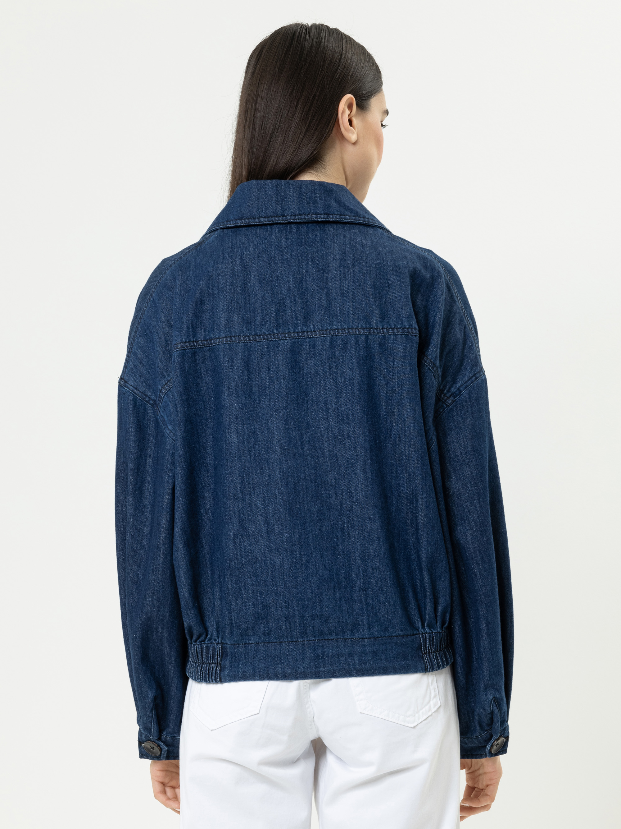 Eine Frau mit glattem dunklem Haar modelliert eine dunkelblaue Jeansjacke, von hinten gesehen, gepaart mit einer weißen Hose vor einem schlichten hellen Hintergrund.