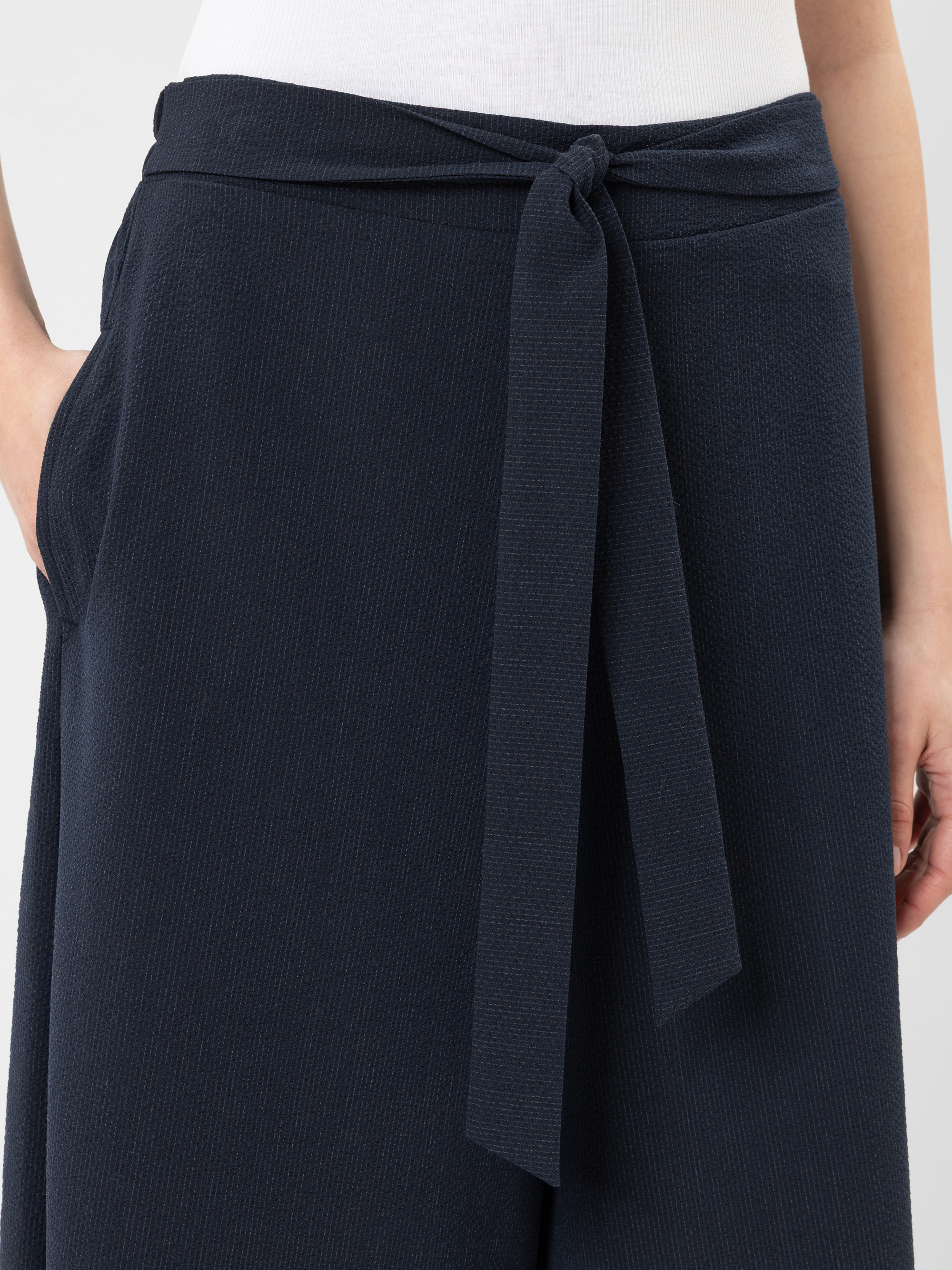 Nahaufnahme einer Person, die eine dunkle marineblaue Hose mit einem Stoffgürtel trägt, der in der Taille verknotet ist. Die Hand der Person steckt in der Tasche, und ein weißes Oberteil ist teilweise sichtbar.