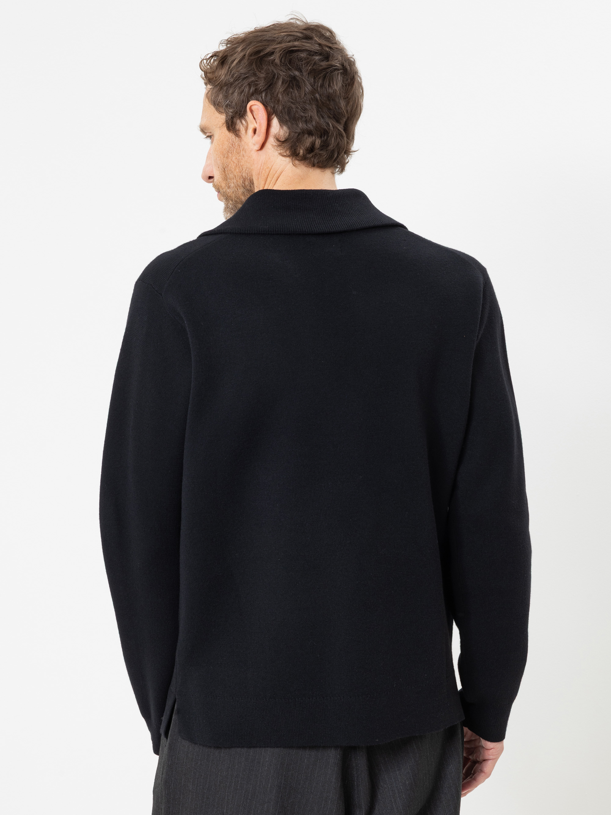 Vor einem schlichten weißen Hintergrund steht ein Mann mit dem Rücken zur Kamera; er trägt einen schwarzen, langärmeligen Pullover mit Kragen und eine dunkle Hose.