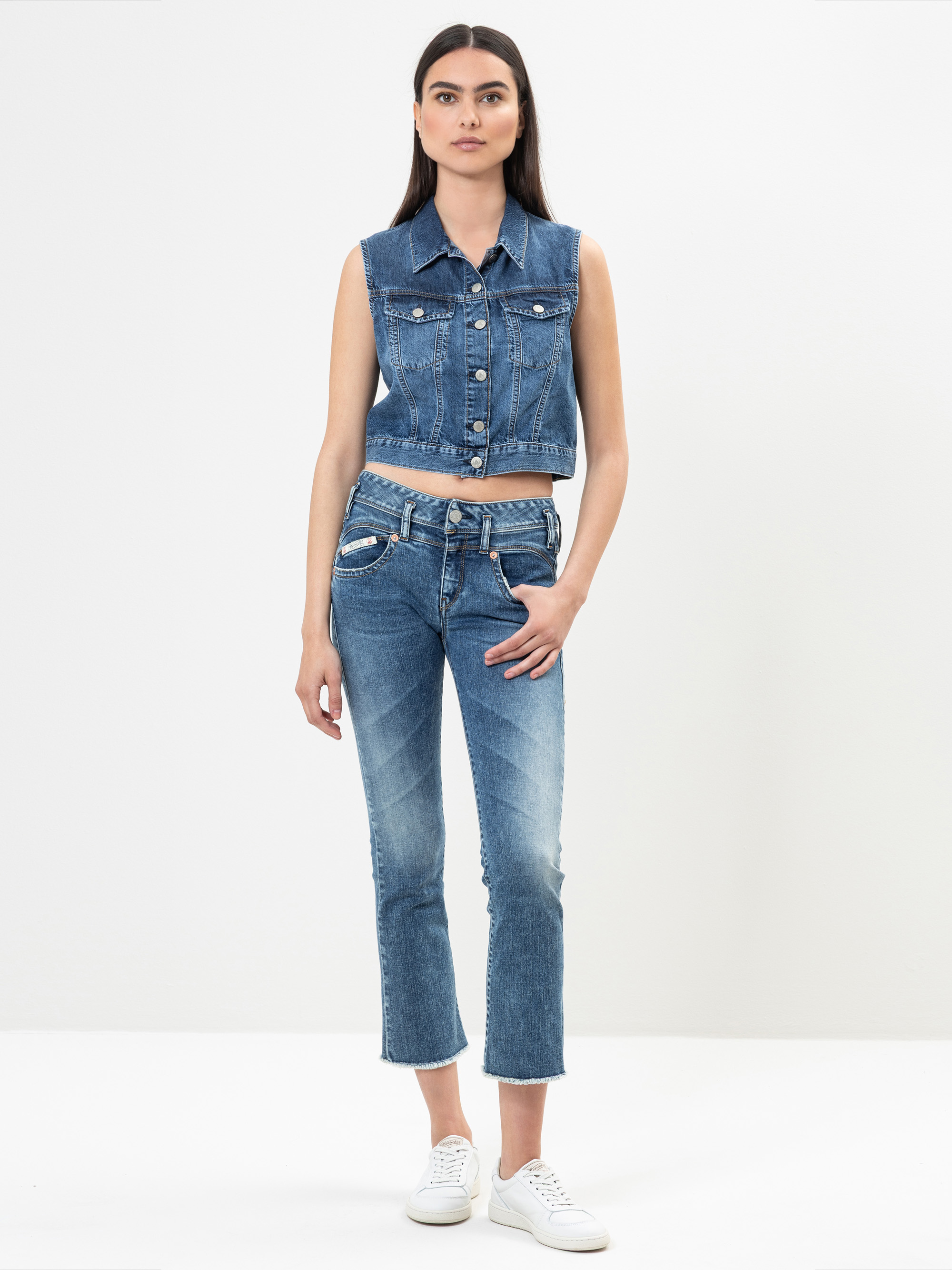 Eine Frau steht vor einem schlichten weißen Hintergrund und trägt eine ärmellose Jeansweste, dazu passende hochgeschnittene Jeans und weiße Turnschuhe. Sie hat langes dunkles Haar und einen neutralen Gesichtsausdruck, eine Hand steckt in ihrer Tasche.