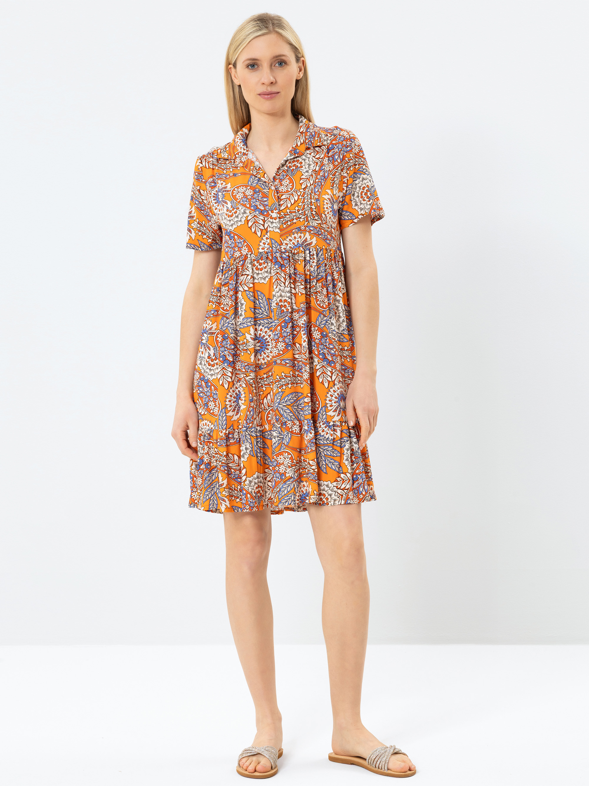 Eine Frau steht vor einem schlichten weißen Hintergrund und trägt ein kurzärmeliges, orange gemustertes Kleid mit Kragen und Sandalen. Sie hat helle Haut, glattes blondes Haar und einen neutralen Gesichtsausdruck.