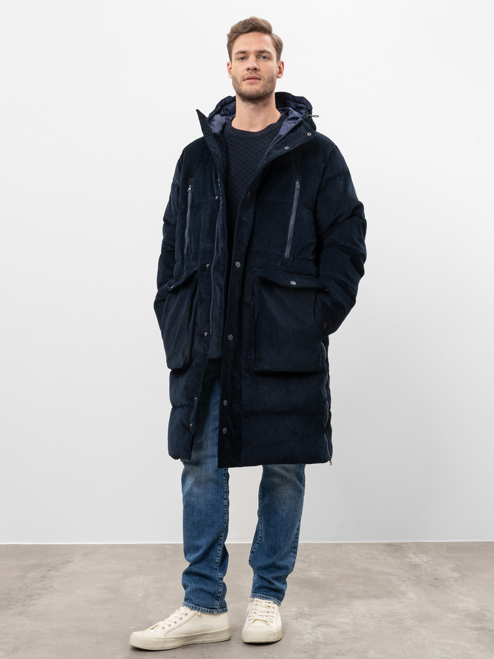 Ein Mann steht mit dem Gesicht nach vorne und trägt einen langen schwarzen Wintermantel, blaue Jeans und weiße Turnschuhe. Der Hintergrund ist einfarbig weiß, und der Boden scheint aus grauem Beton zu bestehen.