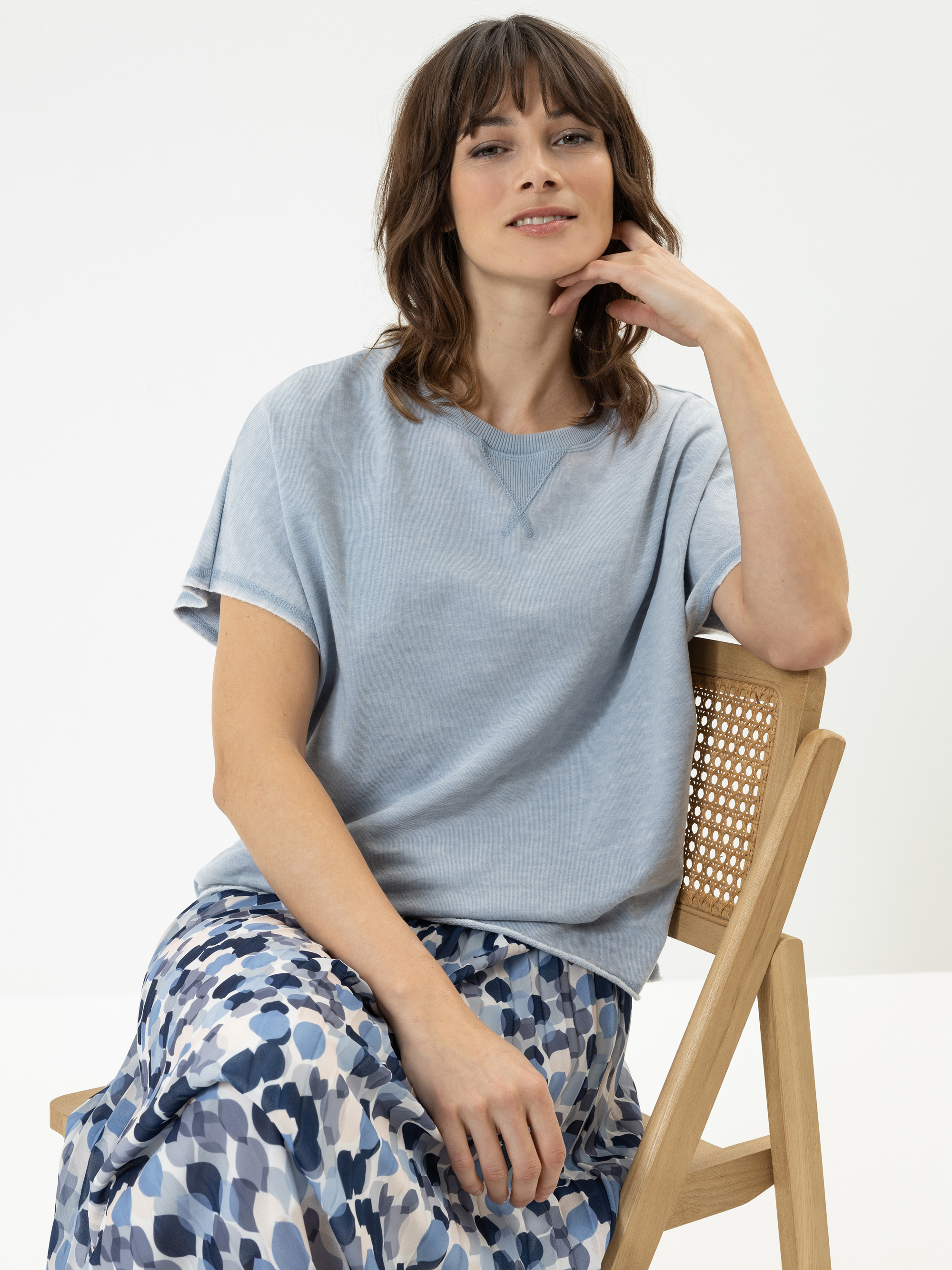 Eine Frau mit schulterlangen braunen Haaren sitzt auf einem Holzstuhl, trägt ein hellblaues kurzärmeliges Oberteil und einen fließenden Rock mit einem blau-weißen abstrakten Muster und posiert mit einem entspannten, selbstbewussten Ausdruck.
