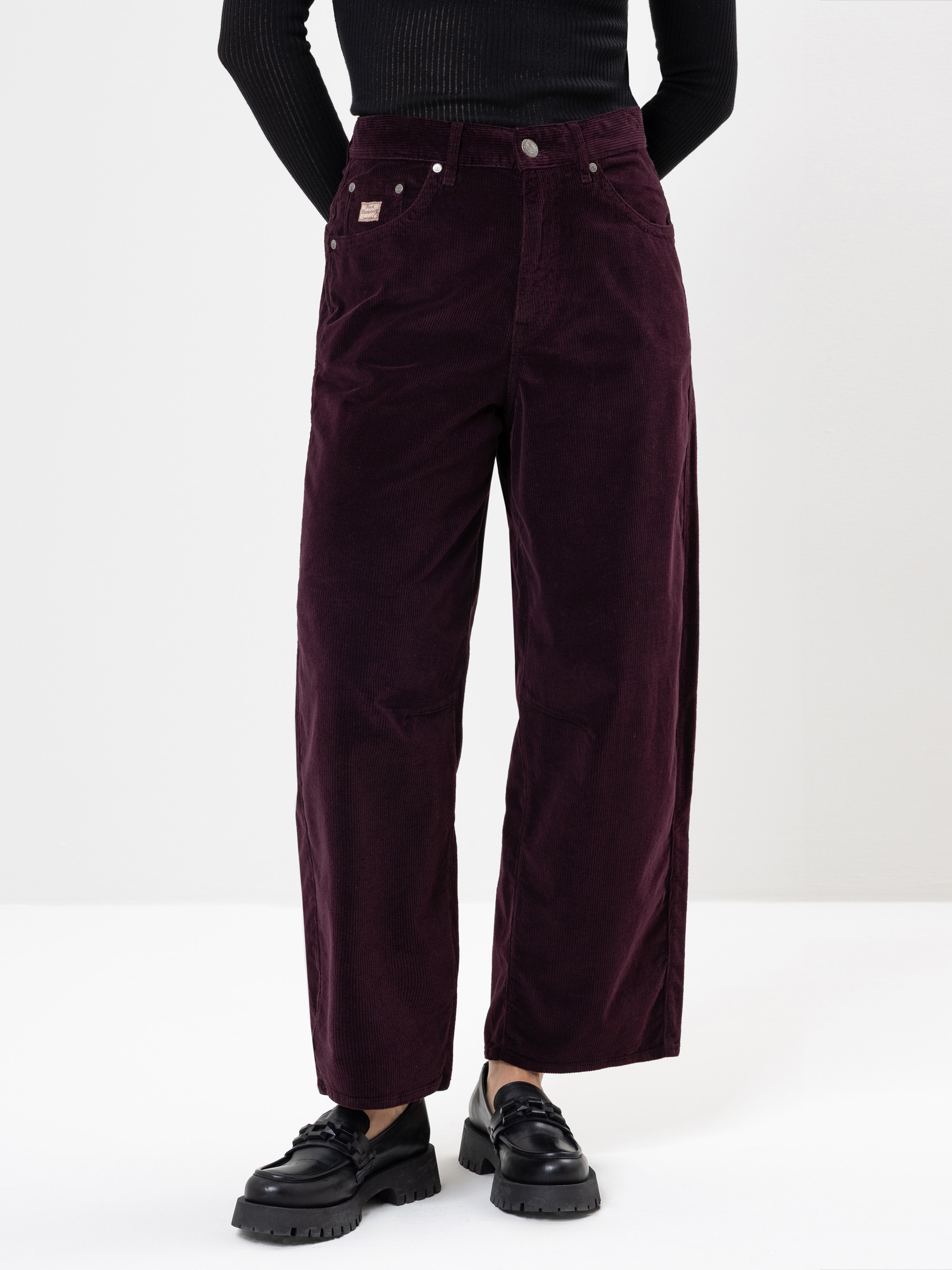 Eine Person, die eine dunkellila Cordhose mit hoher Taille und weitem Bein, ein schwarzes langärmeliges Rippenoberteil und schwarze klobige Halbschuhe trägt, steht vor einem schlichten weißen Hintergrund.