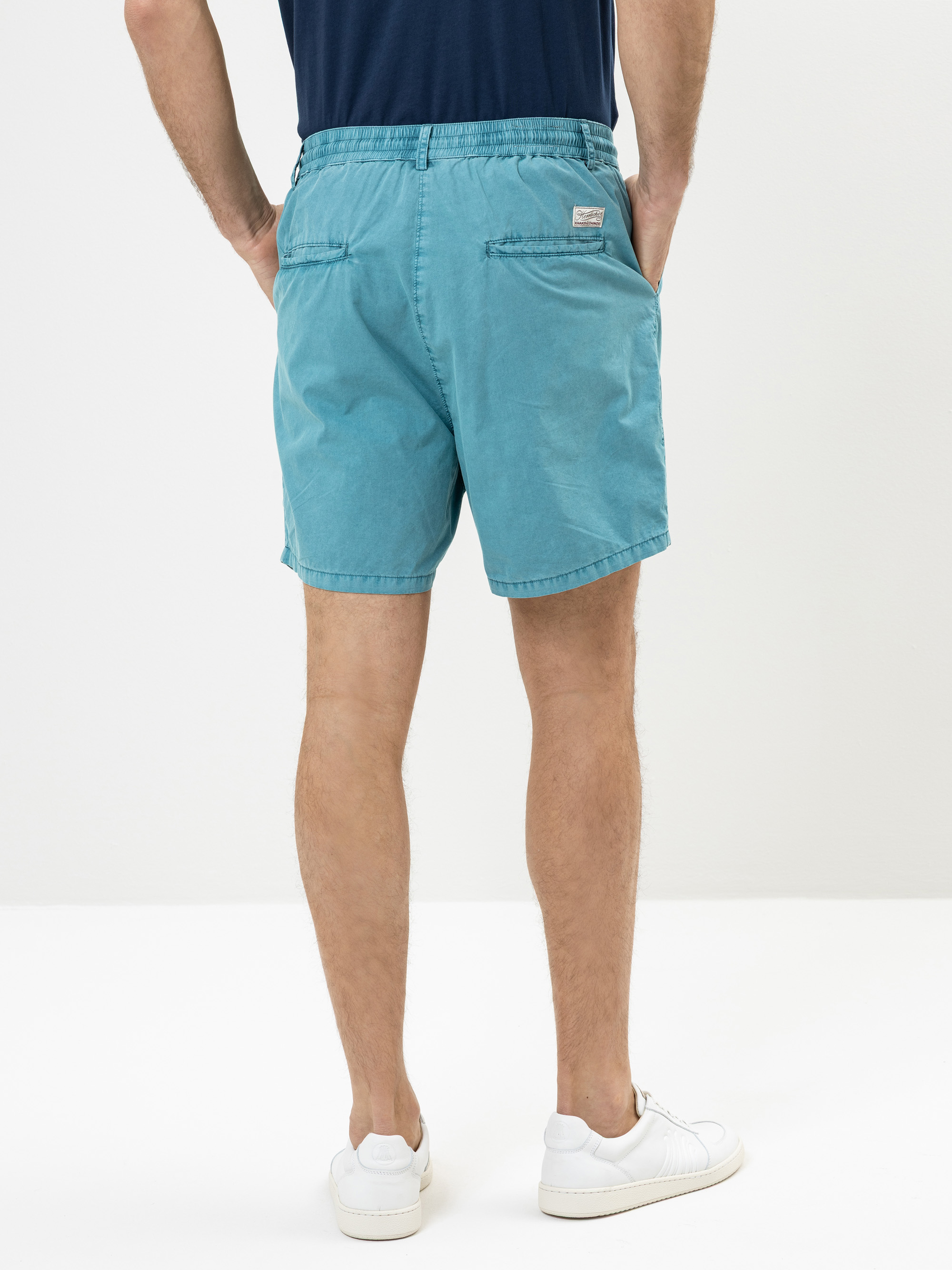 Ein Mann steht mit dem Gesicht zur Wand, trägt blaugrüne Shorts, ein marineblaues T-Shirt und weiße Turnschuhe, die Hände in den Gesäßtaschen, vor einem schlichten weißen Hintergrund.