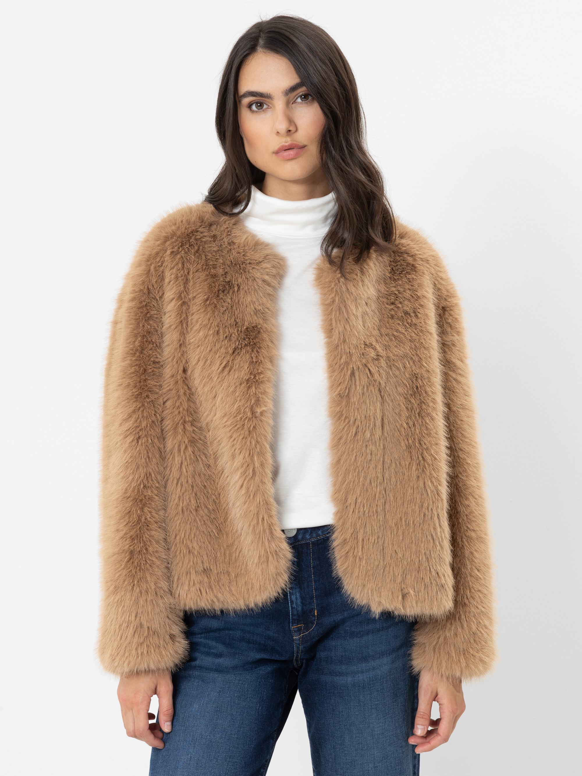 Eine Frau mit langen dunklen Haaren, die eine hellbraune Kunstfelljacke über einem weißen Rollkragenpullover und einer blauen Jeans trägt und vor einem schlichten weißen Hintergrund steht.