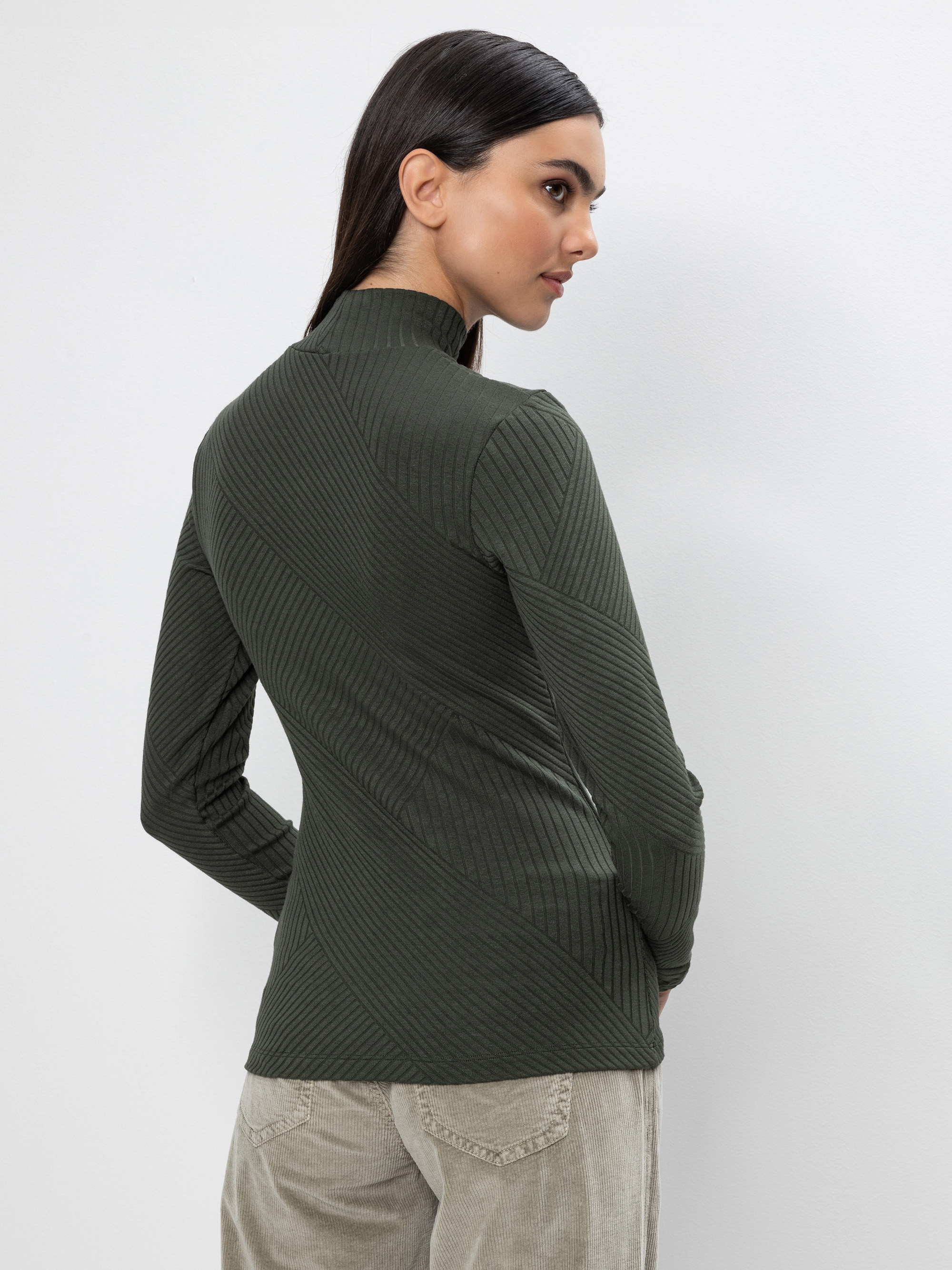Eine Frau mit langen dunklen Haaren steht mit dem Gesicht zur Wand und trägt einen dunkelgrünen, gerippten Rollkragenpullover und eine hellbeige Hose vor einem schlichten hellen Hintergrund.