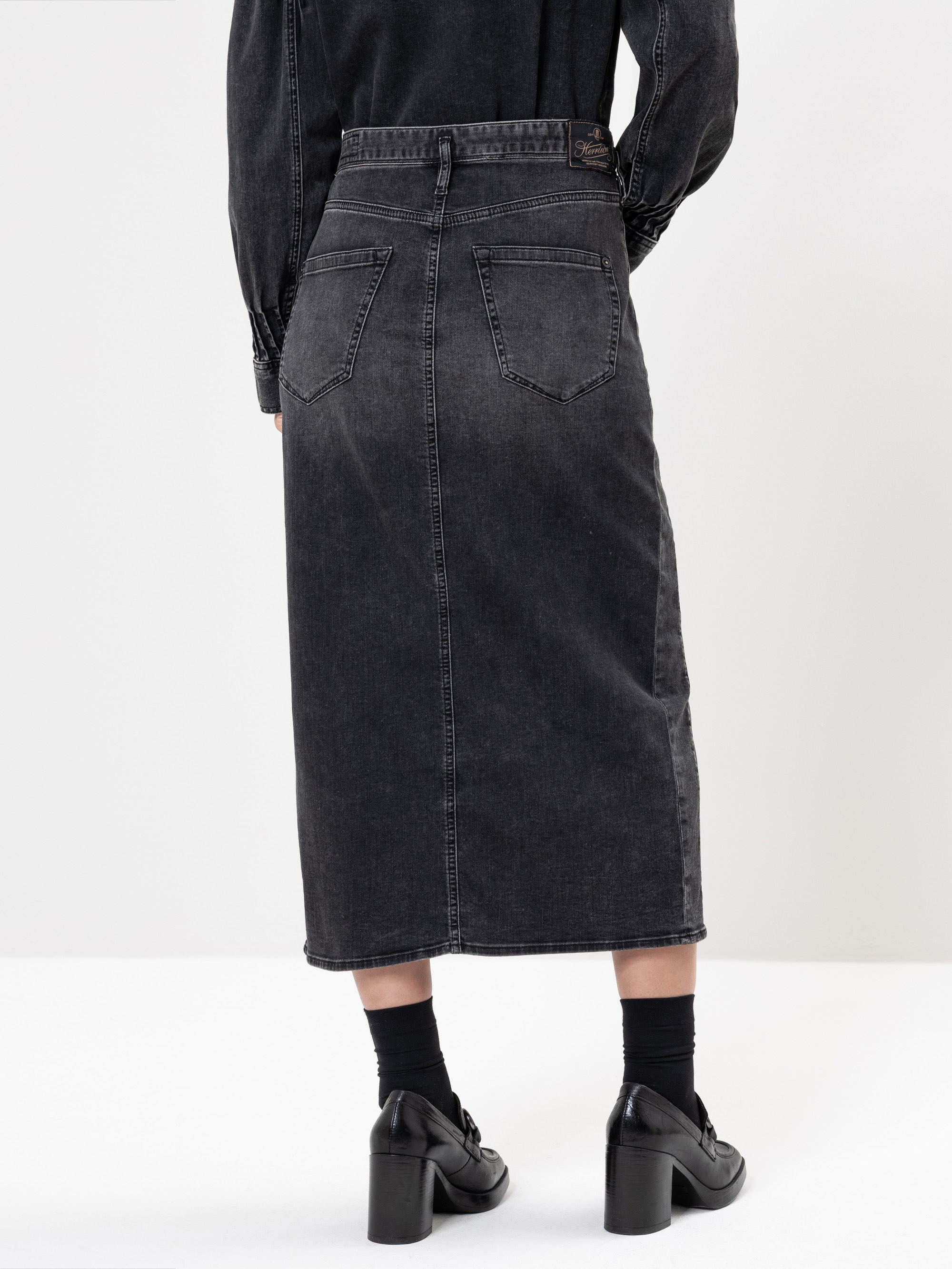 Eine Person, die einen hochtaillierten, schwarzen Jeans-Midirock mit Gesäßtaschen, schwarzen Socken und schwarzen Schuhen mit Absätzen trägt, wird von hinten vor einem schlichten weißen Hintergrund gezeigt.