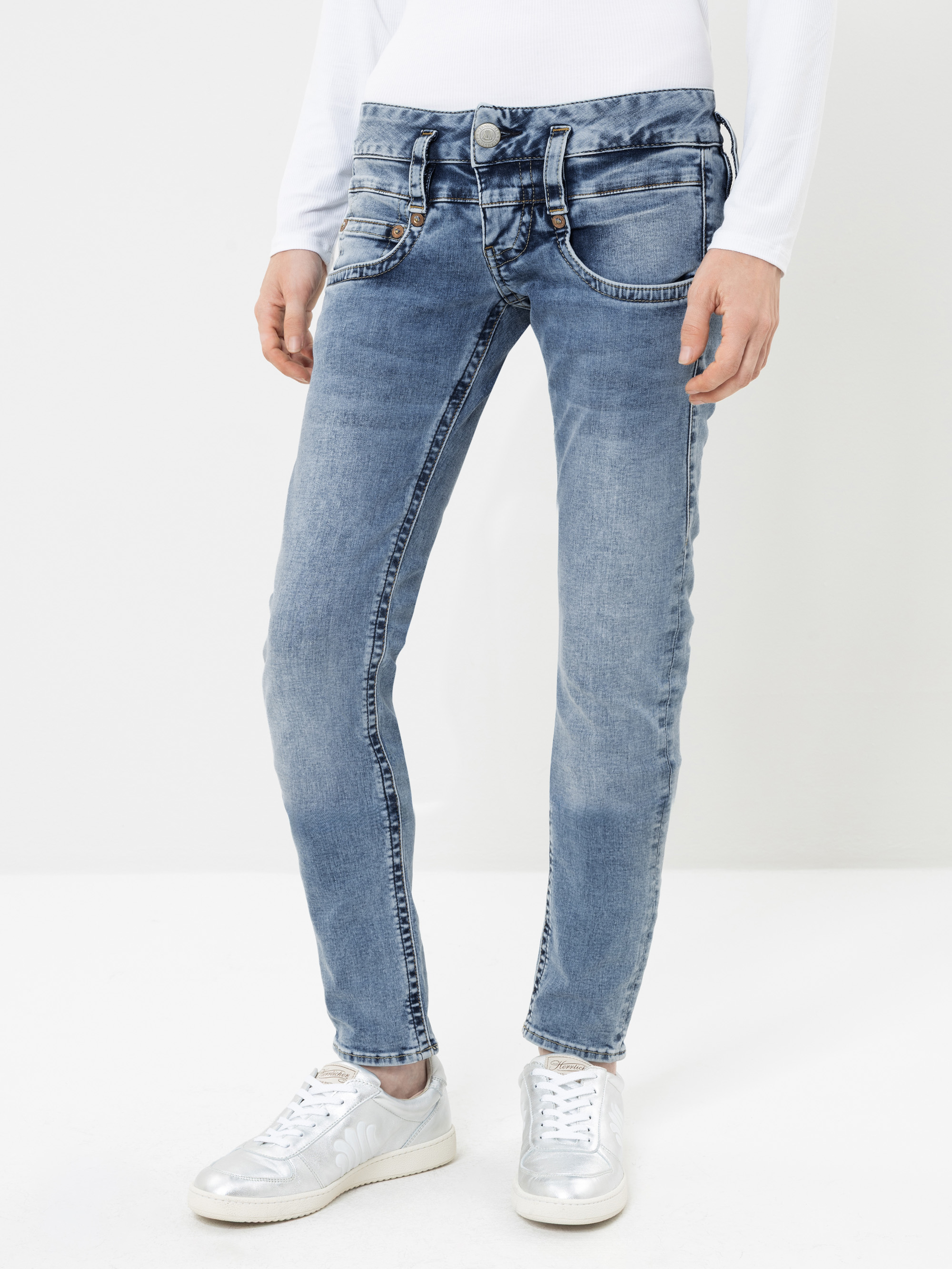 Eine Person, die hellblaue, ausgewaschene Jeans mit mehreren Vordertaschen, ein weißes Langarmhemd und silberfarbene Sneaker trägt, steht vor einem weißen Hintergrund.