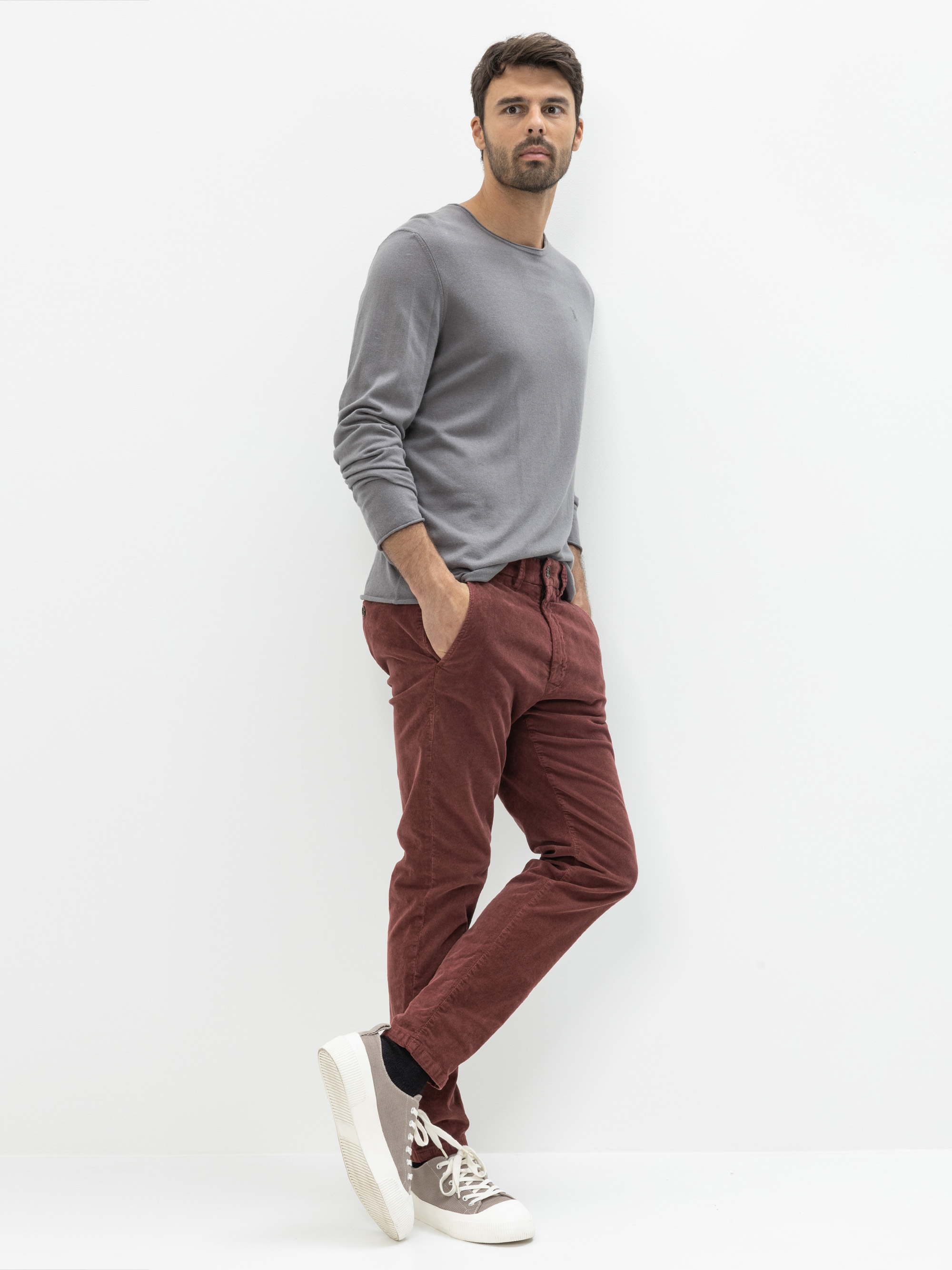 Ein Mann steht vor einem weißen Hintergrund, trägt ein graues, langärmeliges Hemd, eine kastanienbraune Hose, weiße Turnschuhe mit schwarzen Socken und hat die Hände in den Taschen.