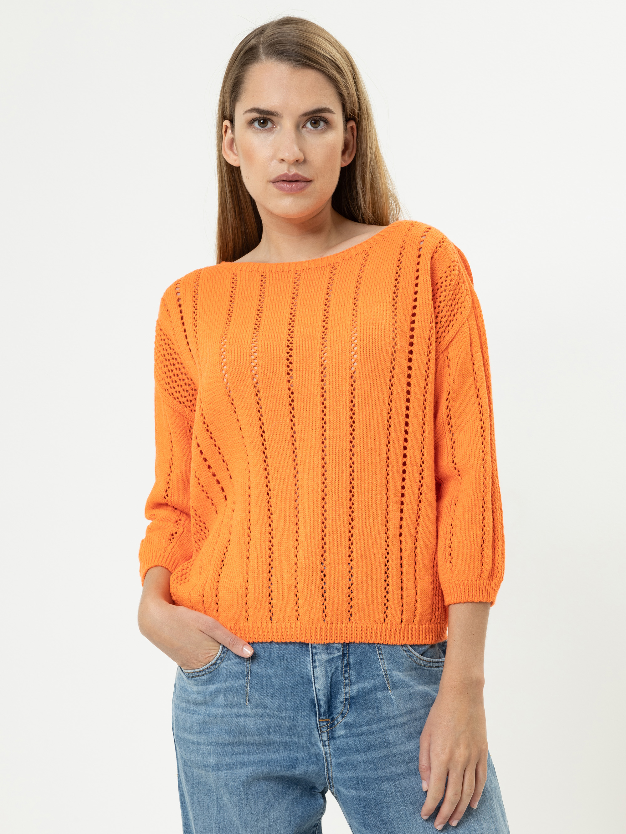Eine Frau mit glattem, hellbraunem Haar trägt einen orangefarbenen Strickpullover mit vertikalen Mustern und blaue Jeans. Sie steht vor einem schlichten weißen Hintergrund, hat eine Hand in der Tasche und schaut in die Kamera.