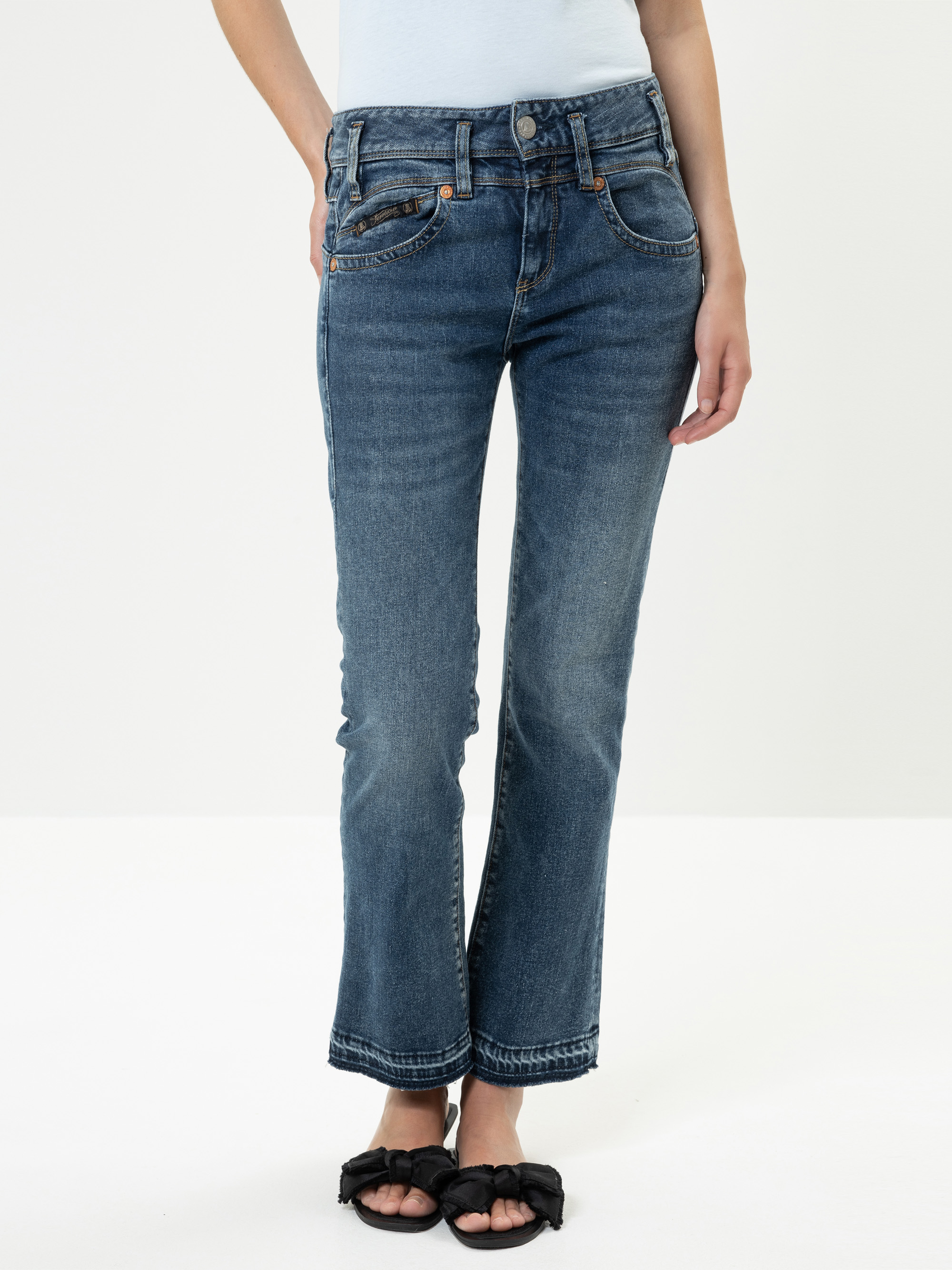 Eine Person, die blaue Jeans mit geradem Bein und schwarze Sandalen trägt, steht vor einem schlichten weißen Hintergrund. Nur der untere Teil des Oberkörpers, die Beine und die Füße sind sichtbar.