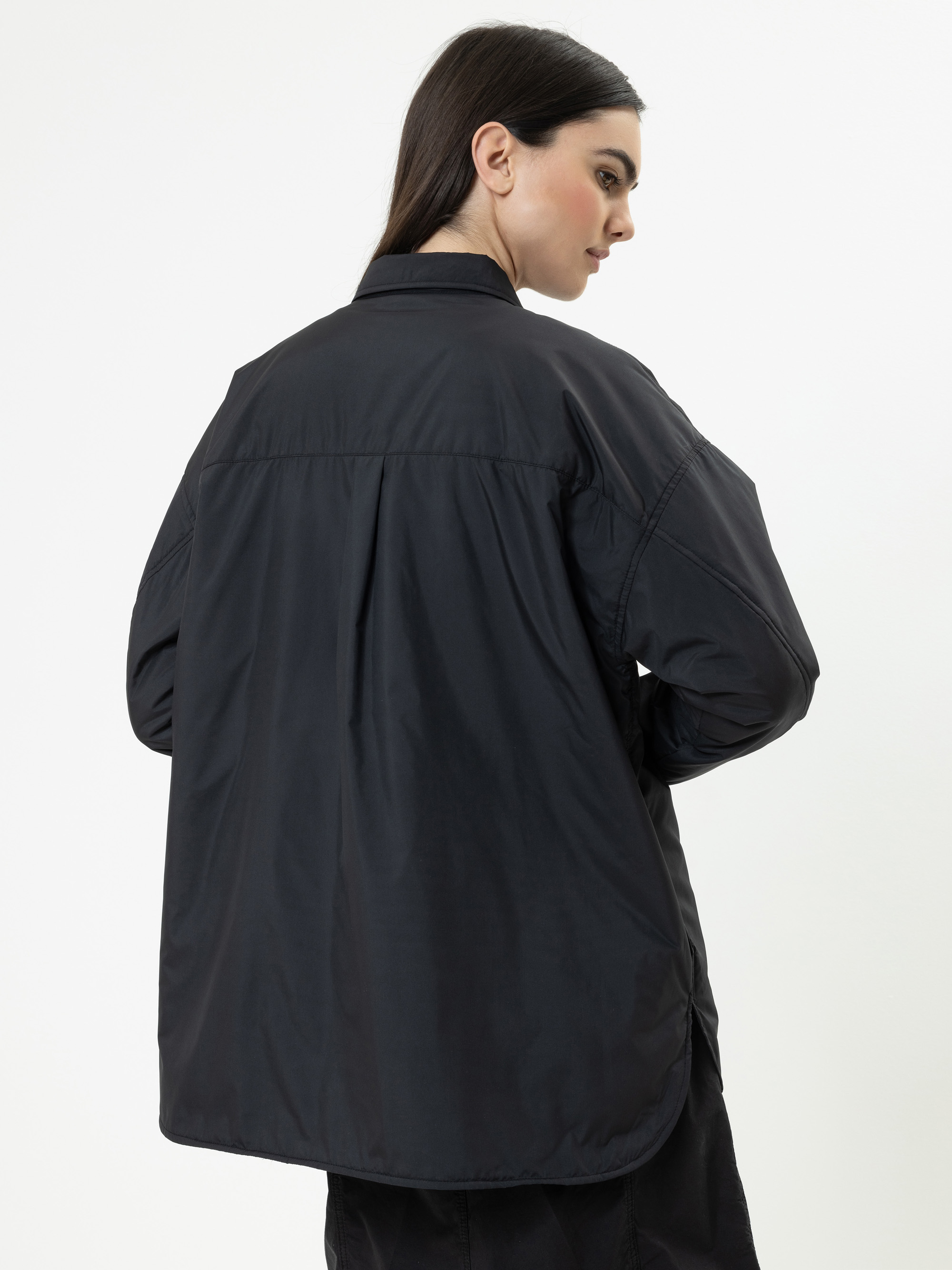 Eine Frau mit langen dunklen Haaren, die eine weite schwarze Jacke trägt, steht mit dem Gesicht zur Seite und blickt über ihre Schulter auf einen schlichten weißen Hintergrund.