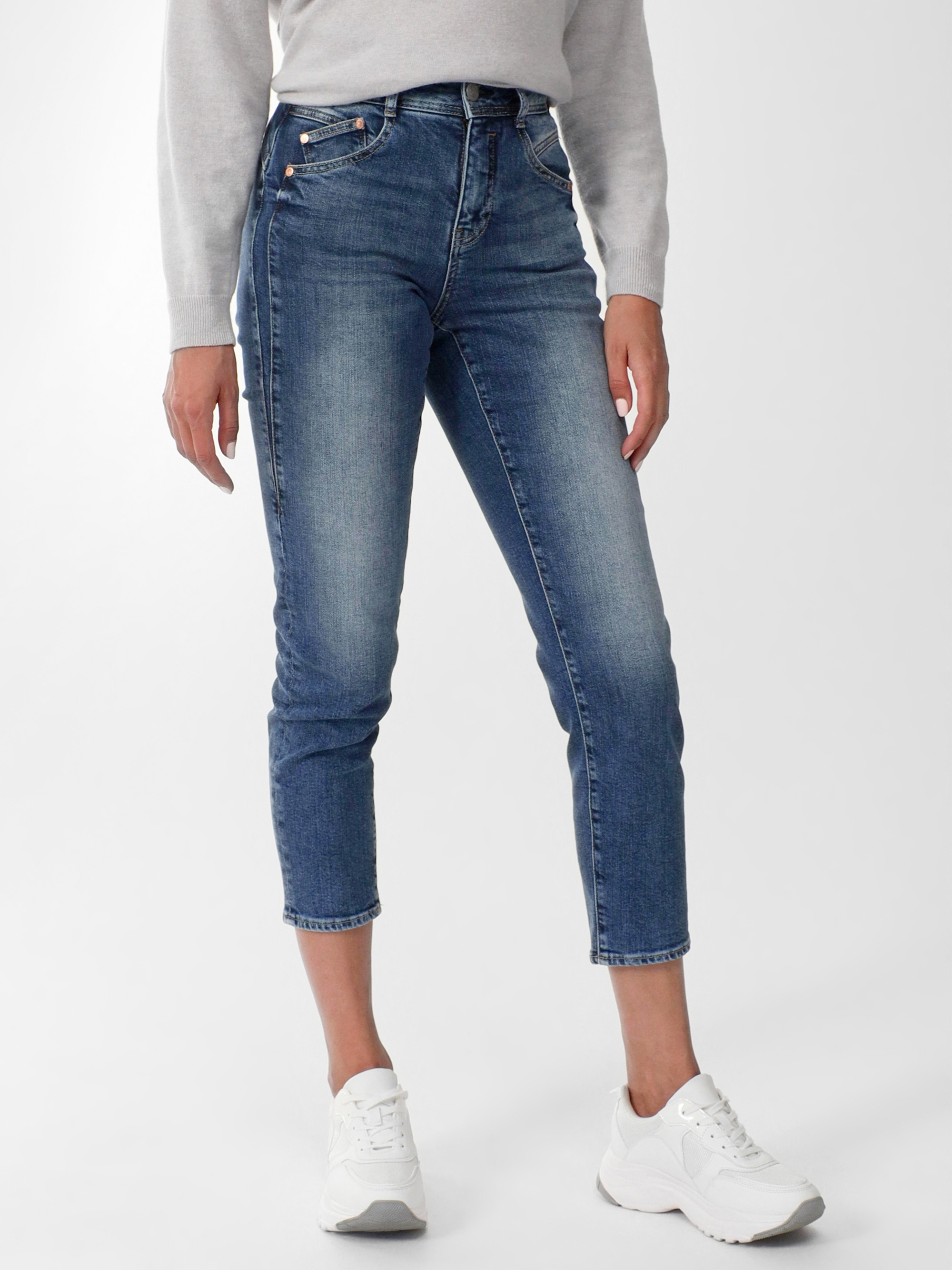 Eine Person, die einen hellgrauen Pullover, blaue Röhrenjeans und weiße Turnschuhe trägt, steht vor einem schlichten weißen Hintergrund. Die Hände der Person liegen entspannt an der Seite.